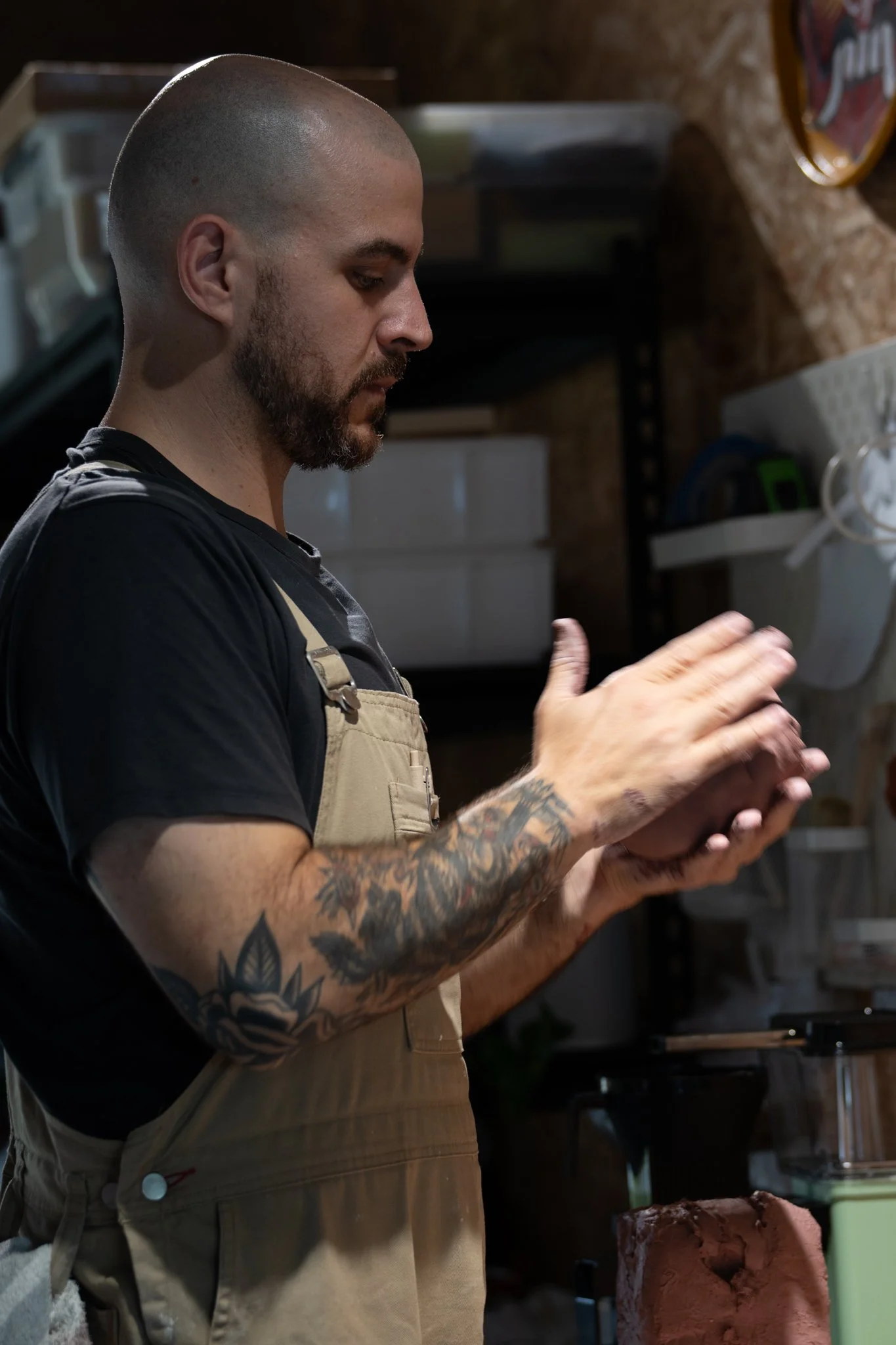 A man with tattoos on his left arm and beard, wearing a black shirt and beige apron, is focused on a task in a kitchen setting. The background shows shelves, containers, and a cake in front of him.