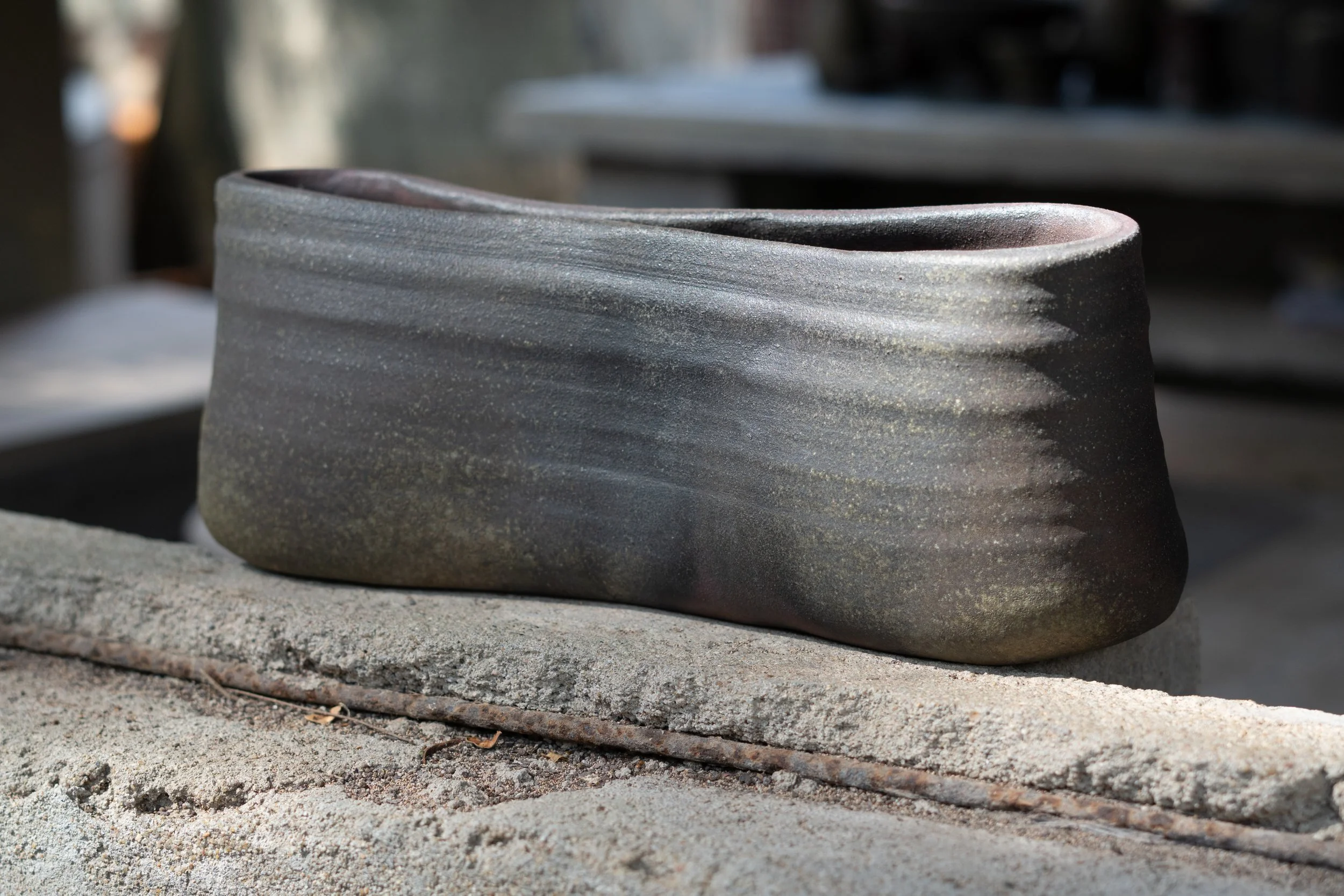 A pottery vase with a wavy texture sits on a stone surface outdoors, with a blurred background.