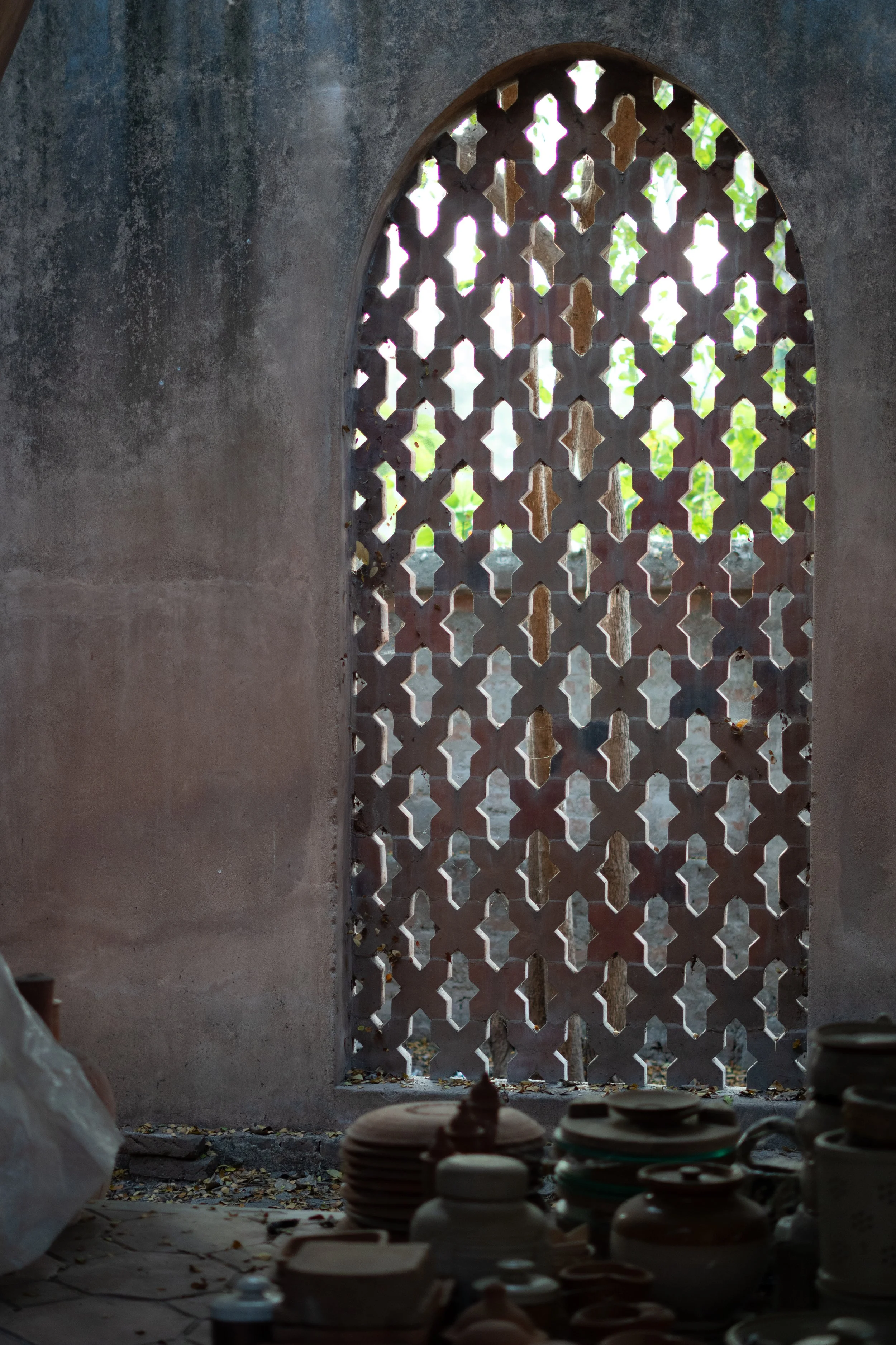 A view through an arched decorative concrete window with geometric lattice pattern, showing a green outdoor scene with trees. Inside, shelves with pottery and dishes are in the foreground, with fallen leaves on the ground.