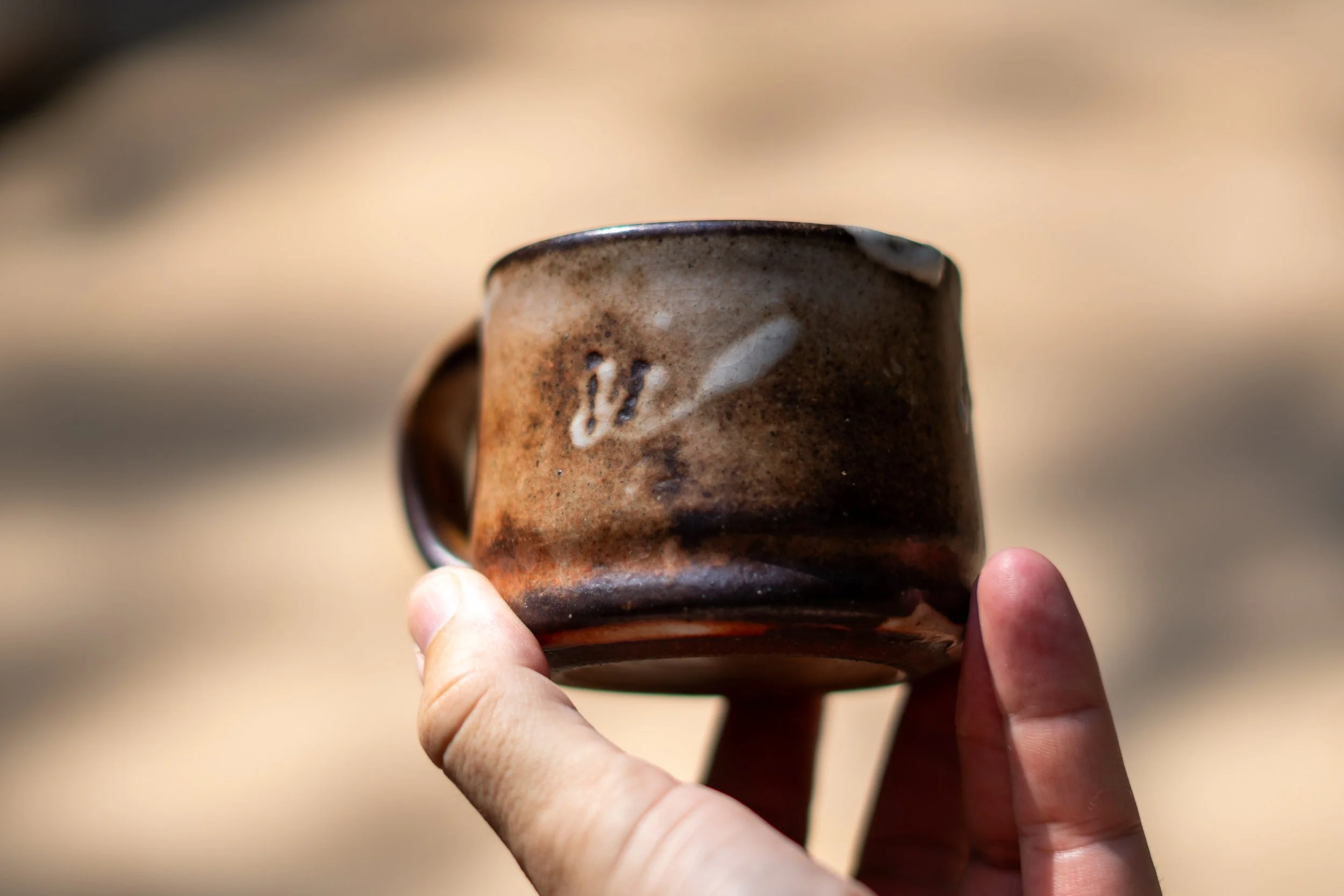 Hand holding a small, rustic brown ceramic mug with a handle, against a blurred neutral background.
