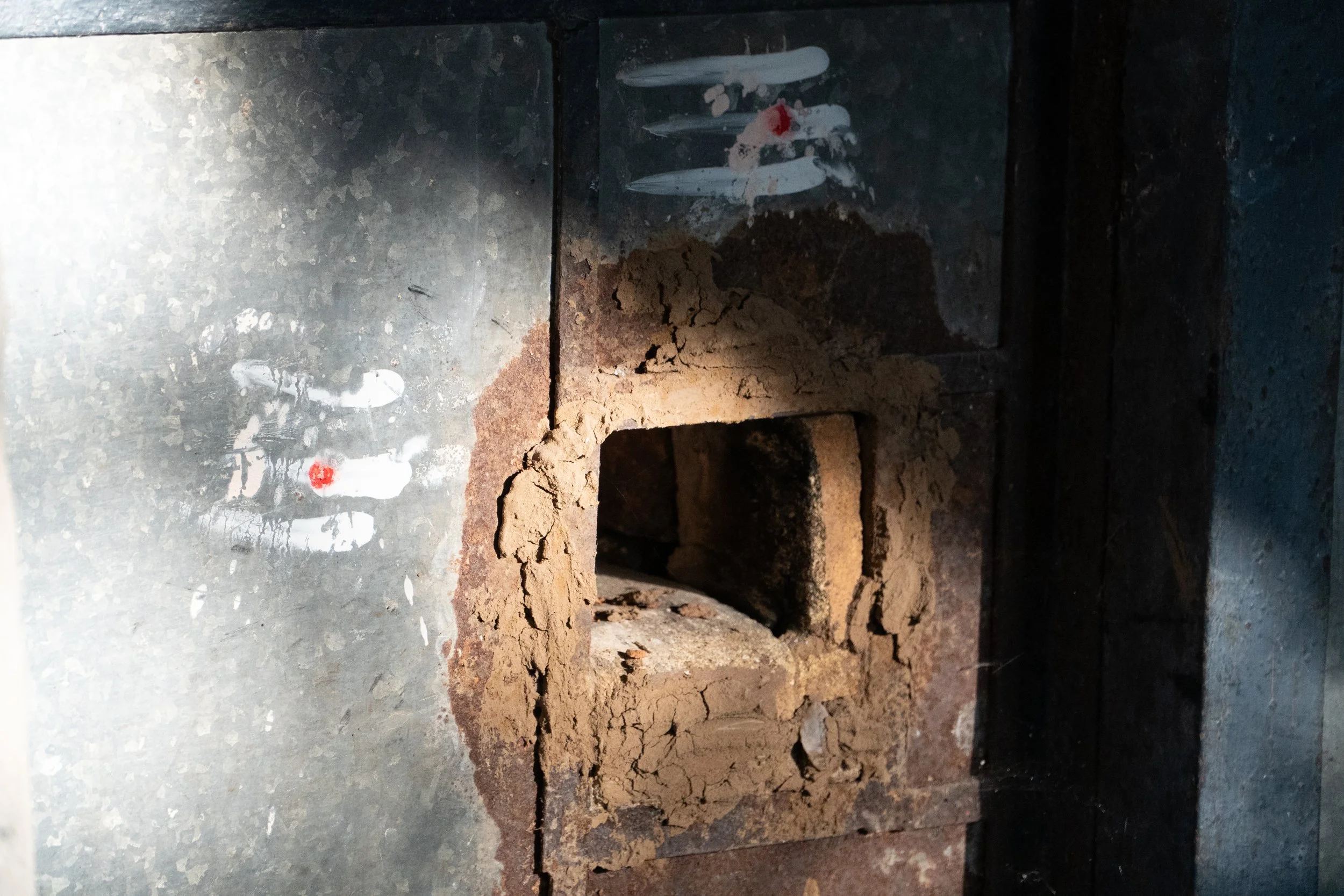 Close-up of a metal surface with paint splatters, dirt, and debris, including broken white fragments and a small red dot, alongside a rough, see-through section revealing dirt and bricks behind.