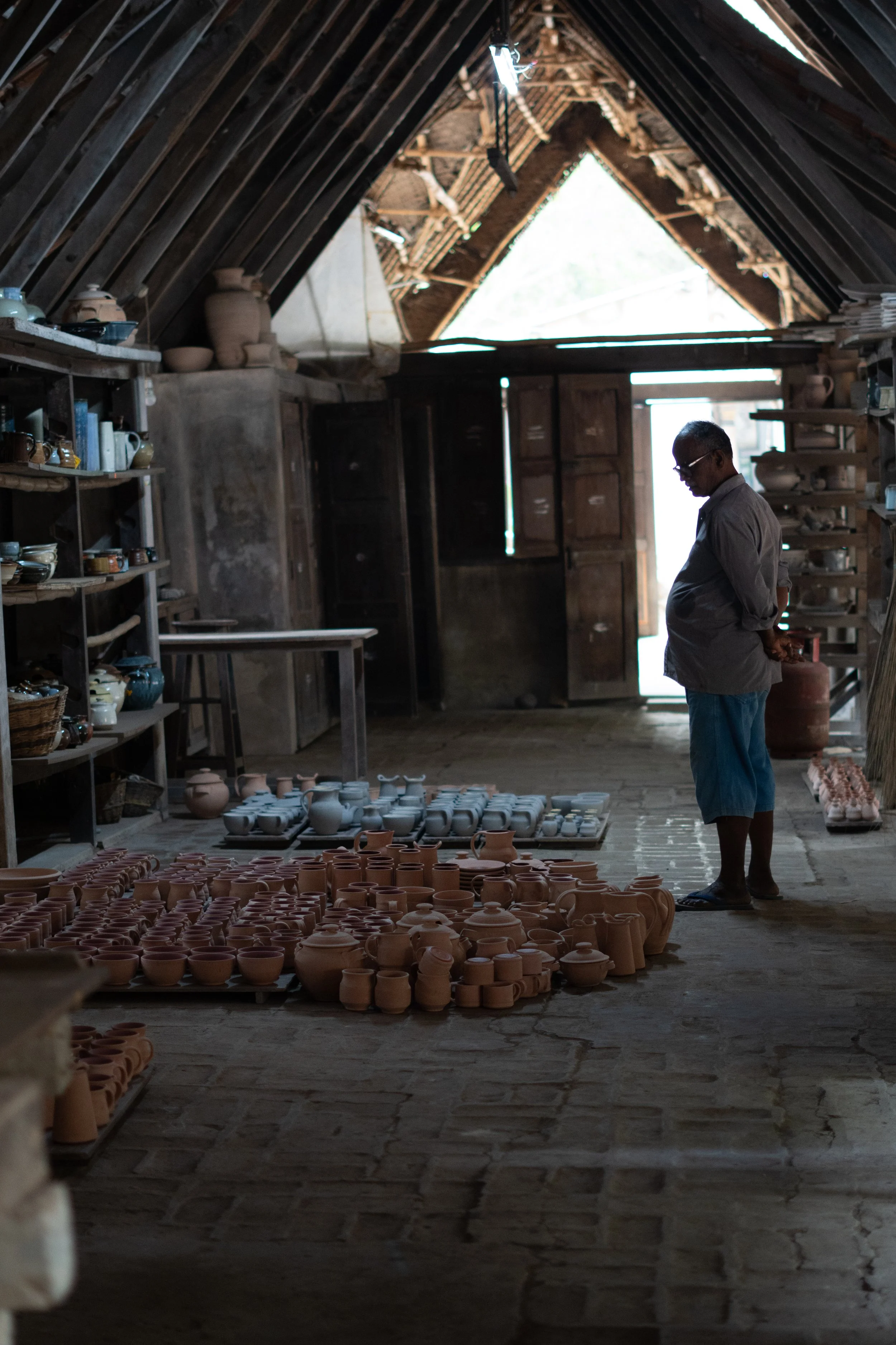 A man observing rows of unglazed pottery in a rustic workshop with shelves of finished pottery, wooden doors, and an open roof allowing natural light.