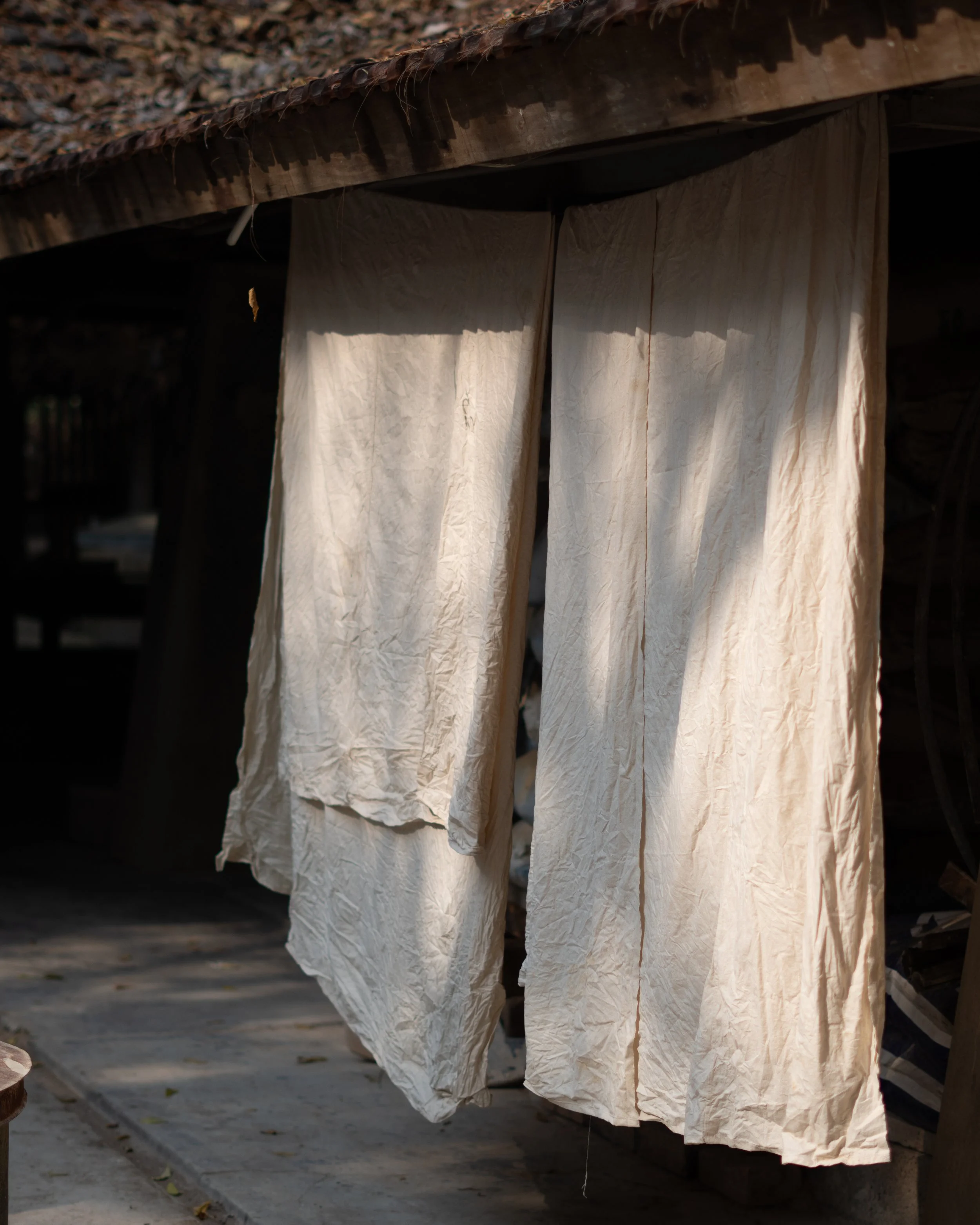 Cream or beige fabric curtains hanging from the edge of a wooden roof with brown shingles, outdoors on a sunny day.