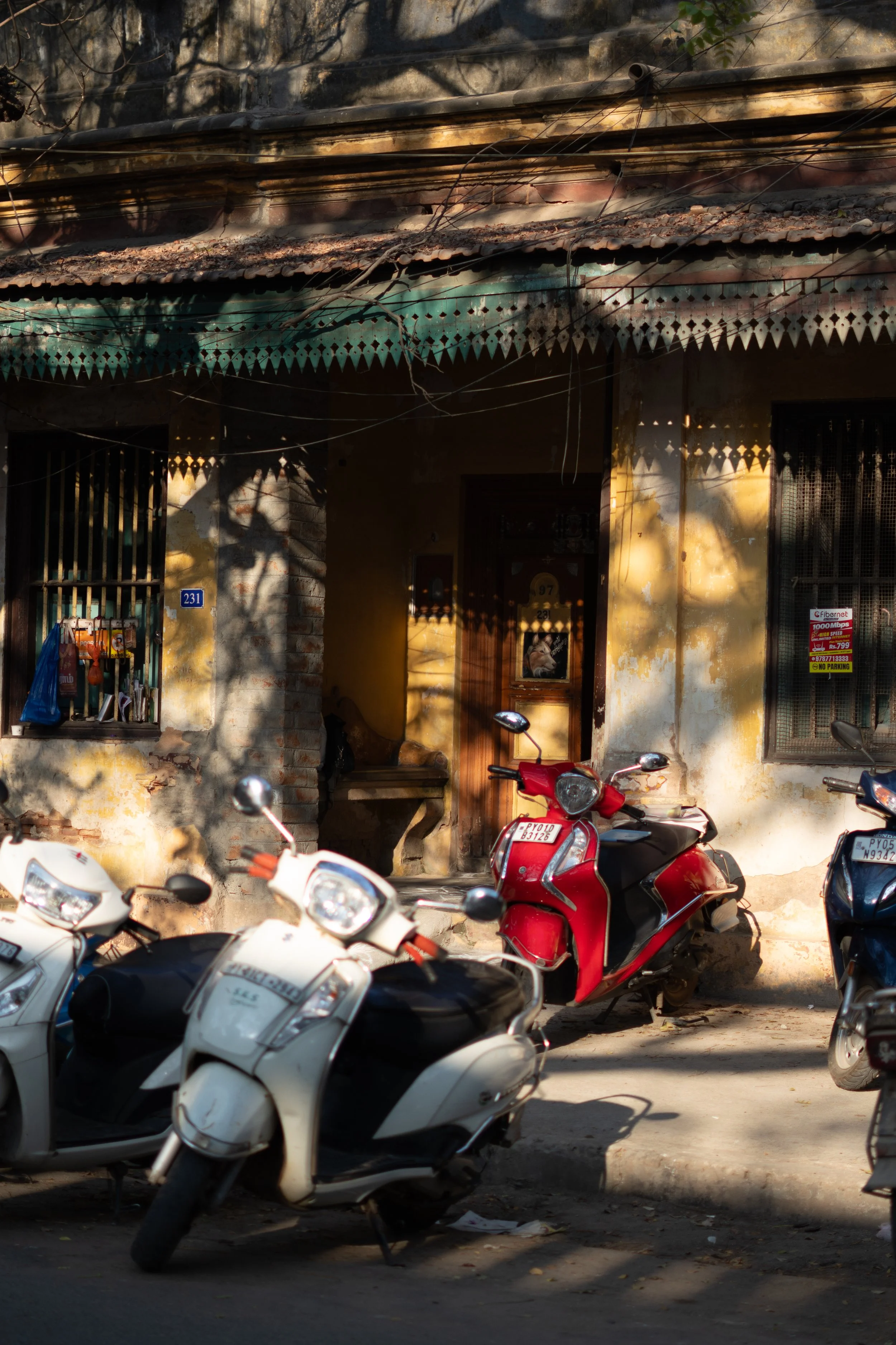 Several parked scooters and motorcycles in front of an old building with a weathered yellow wall, window bars, and a door. Shadows of tree branches cast patterns on the building and motorcycles.