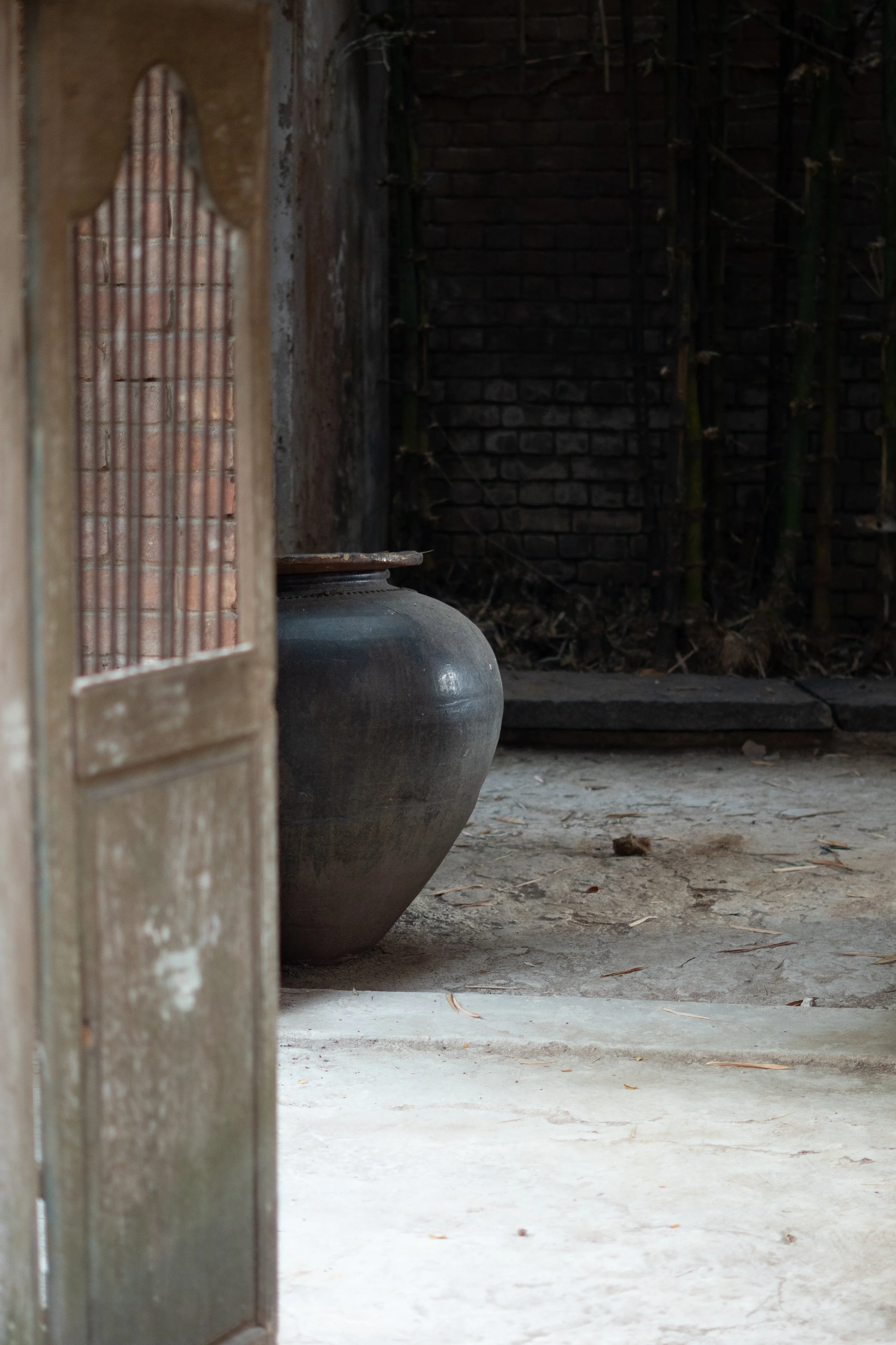 A dark, rustic outdoor area with a brick wall, bamboo stalks, a large ceramic pot, and a wooden door with a decorative window.