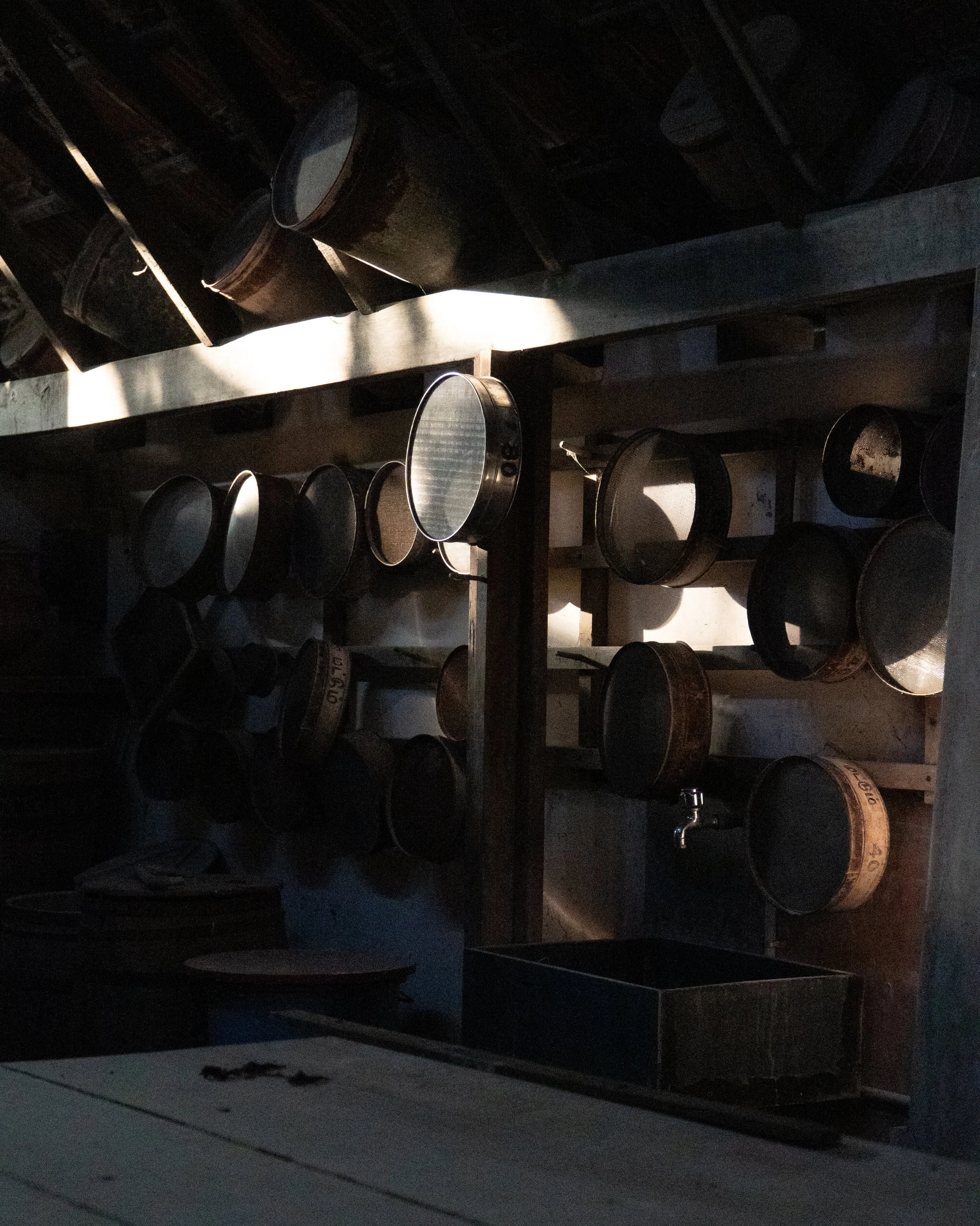 A dimly lit storage room with shelves holding empty black metal containers and some lids, with a box or crate visible at the bottom.
