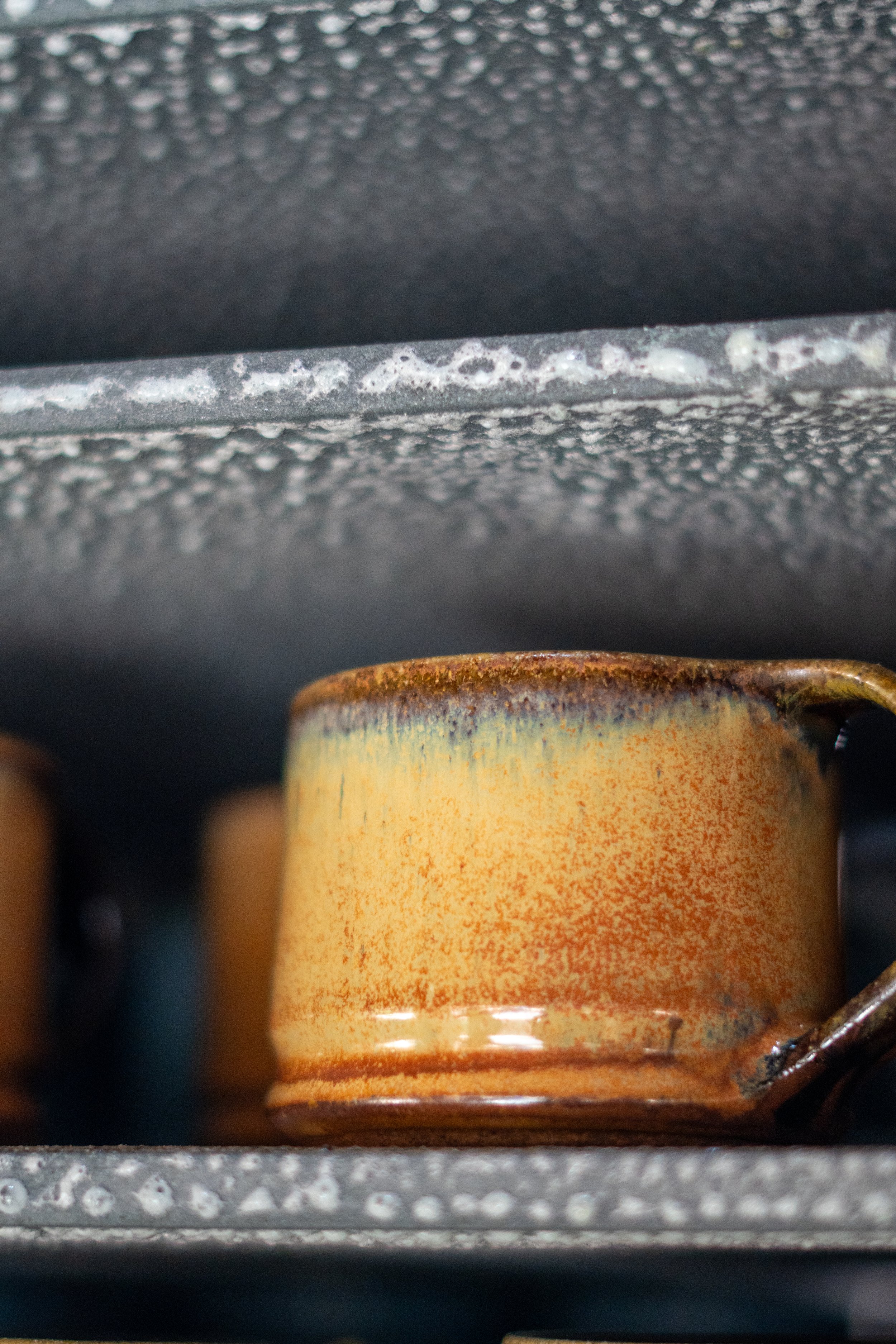 Close-up of a ceramic cup with a gradient glaze of yellow and orange, placed on a metal grate with a rusty surface below.