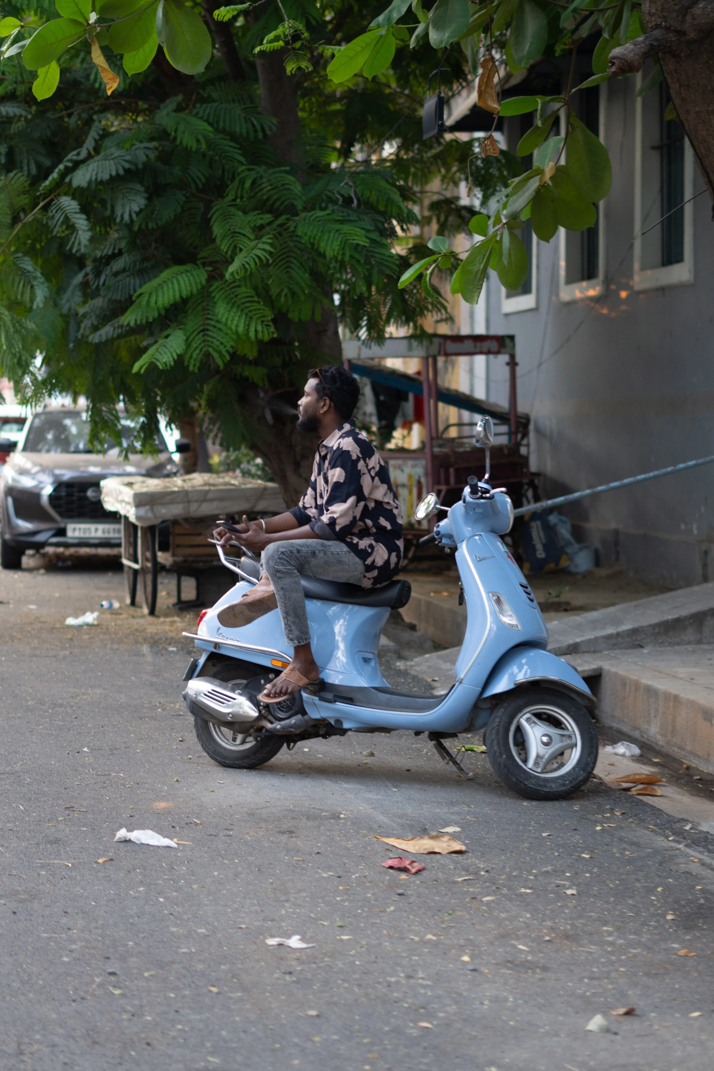 A man with a beard sitting on a light blue scooter parked on the side of the street. He is wearing a patterned shirt and gray pants, holding a phone in his hand. Behind him, there are two cars, a tree with large green leaves, and a building with open