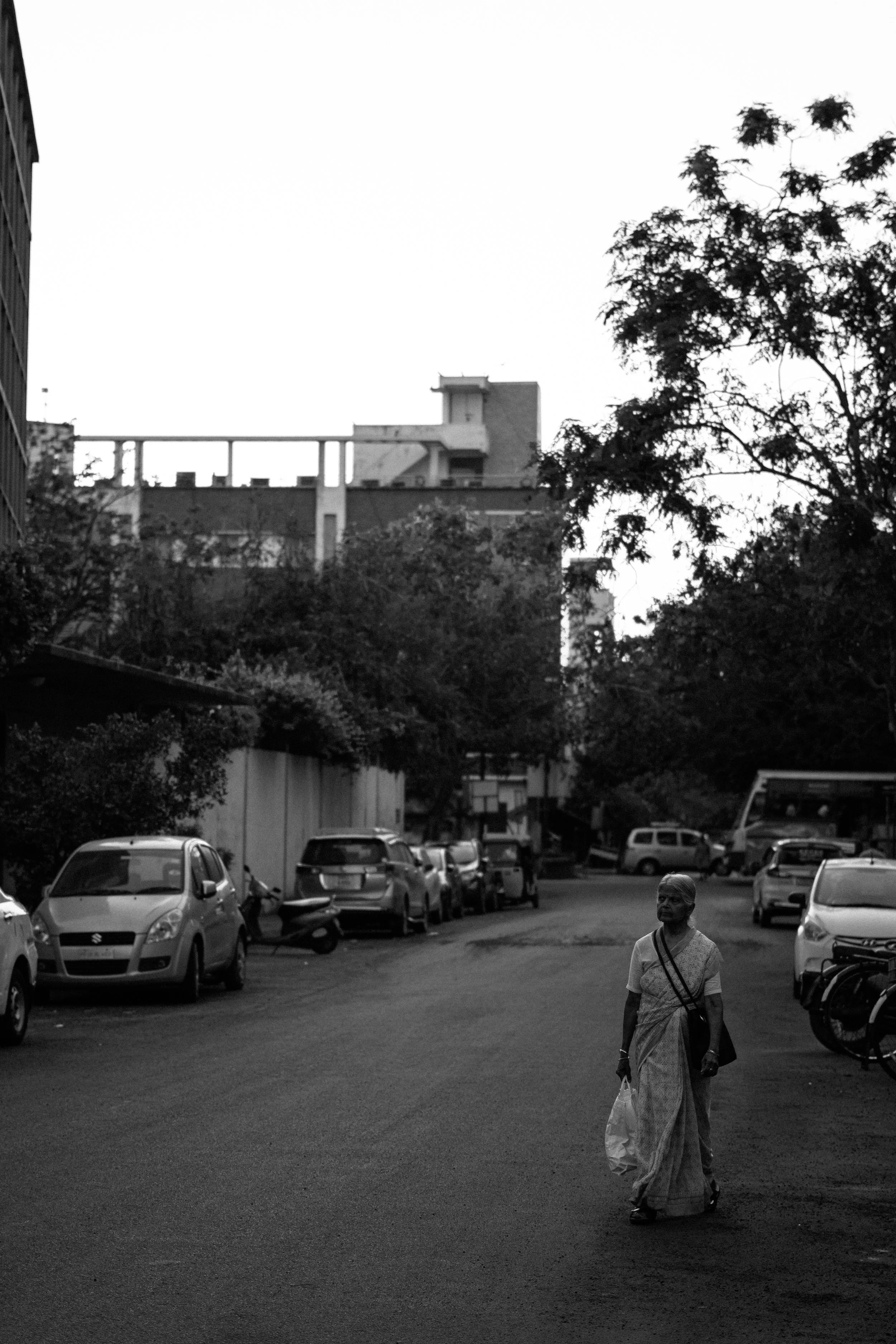 A woman dressed in traditional Indian saree walking on a street with parked cars and trees, with a large building visible in the background.