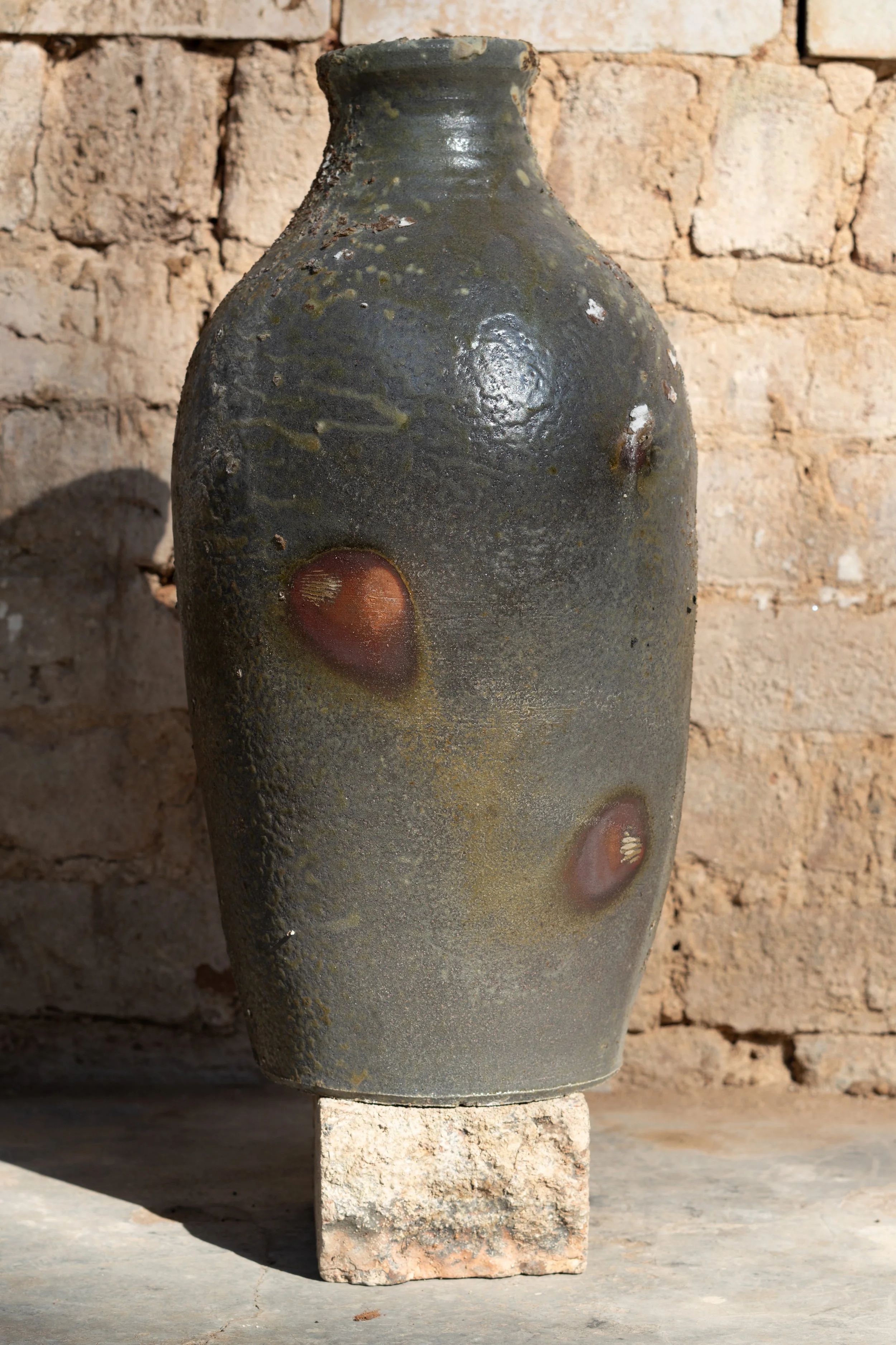 A large, rustic ceramic vase with a dark, textured surface and circular decorative elements, placed on a stone block against a beige brick wall.