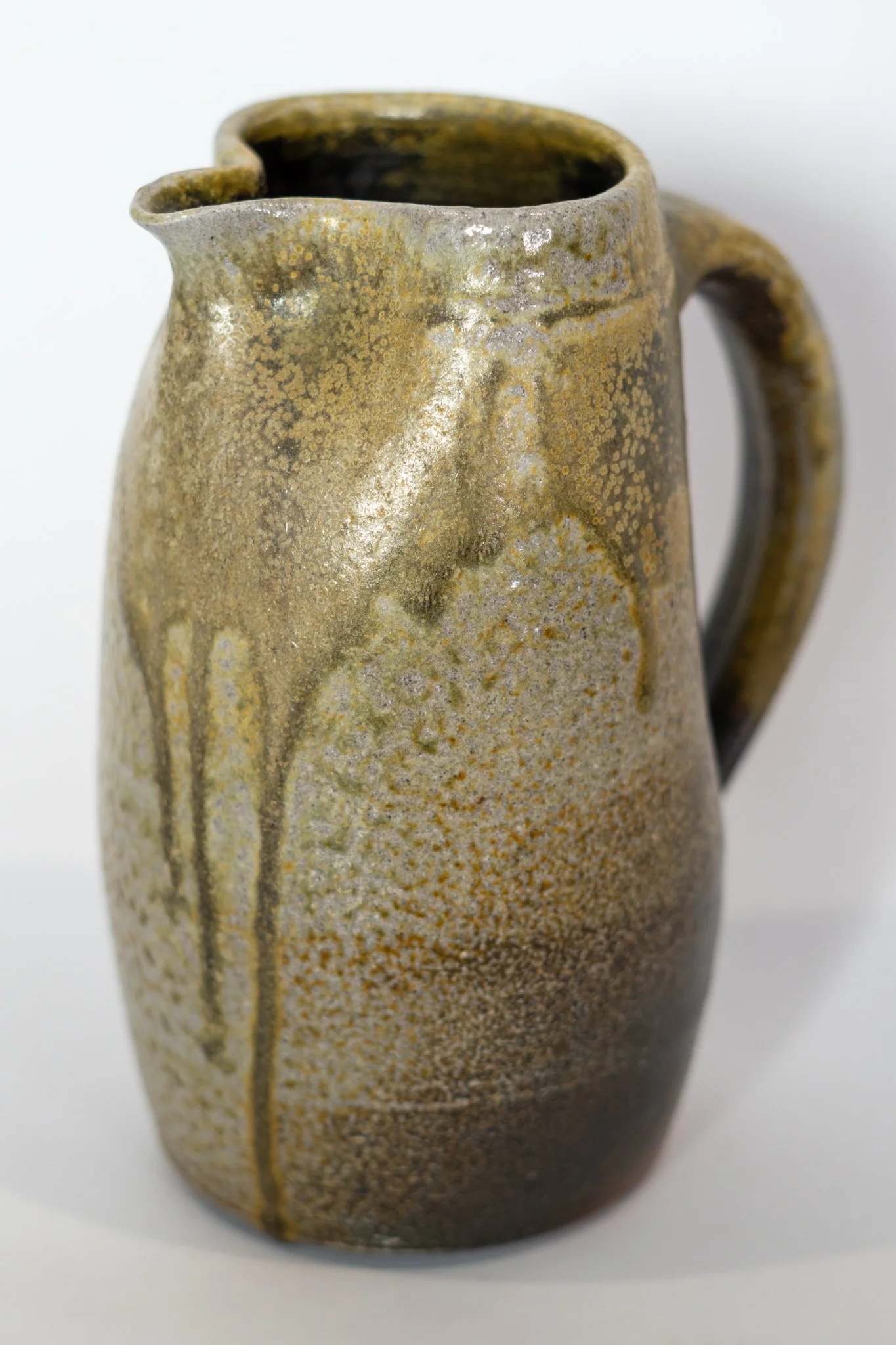 A ceramic pitcher with a textured, speckled glaze finish in earthy tones, featuring a rounded handle and an uneven, slightly chipped lip.
