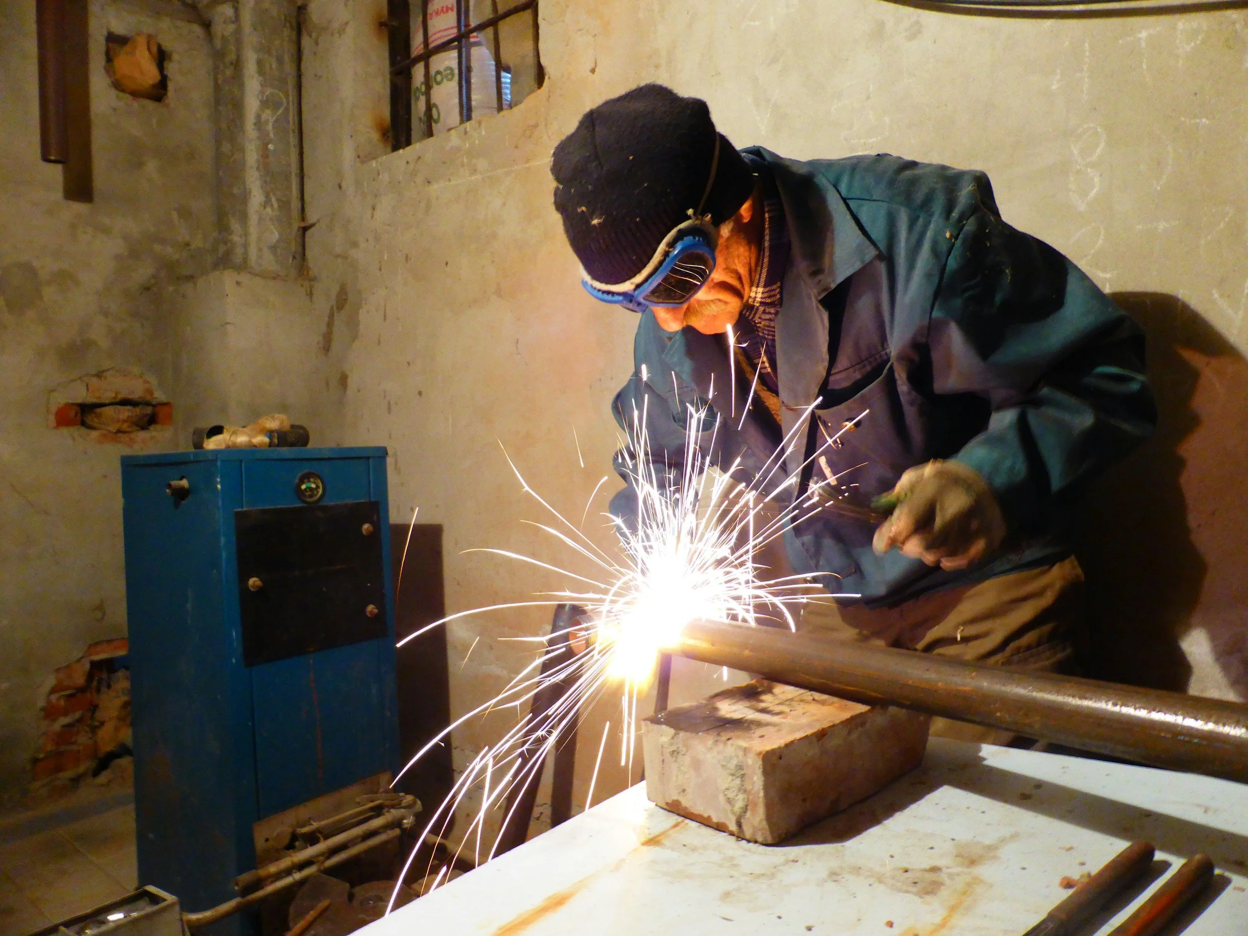 A person welding with protective goggles and gloves in a workshop with a concrete wall and tools around.