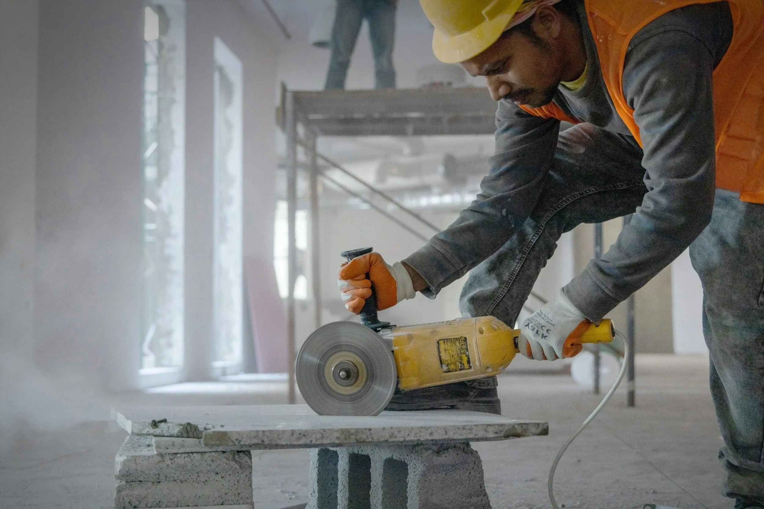 A construction worker wearing a yellow hard hat, an orange safety vest, and gloves uses a handheld circular saw to cut a stone or concrete slab in an industrial setting.