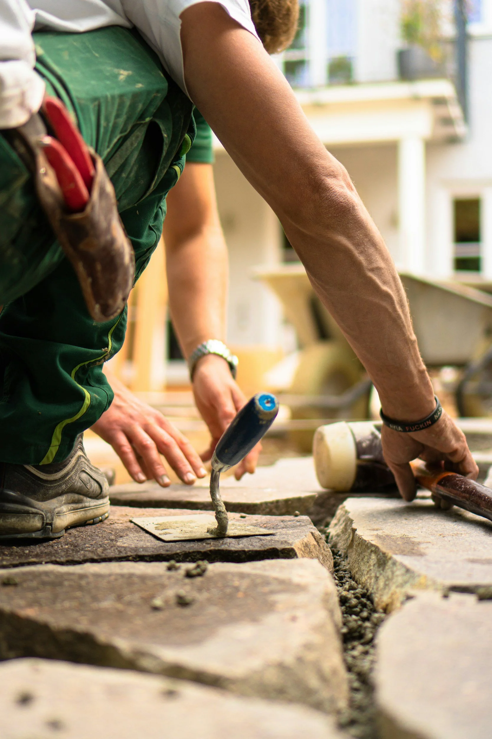 Close-up of construction workers laying bricks on a sidewalk. The focus is on their hands and tools, with one worker's arm extended and another worker helping to position a brick.