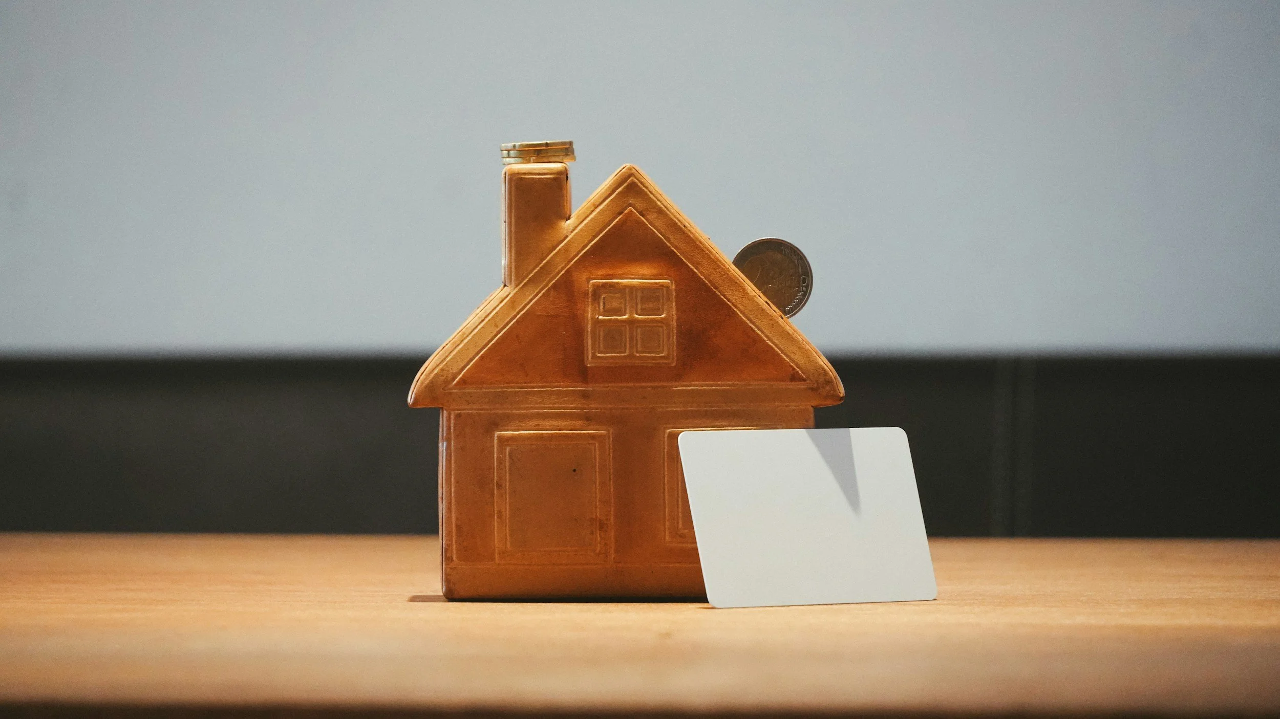 A small house-shaped ceramic container with a chimney, a window, and a door, placed on a wooden surface with a white card standing beside it. Coins are stacked on top of the chimney and behind the container.
