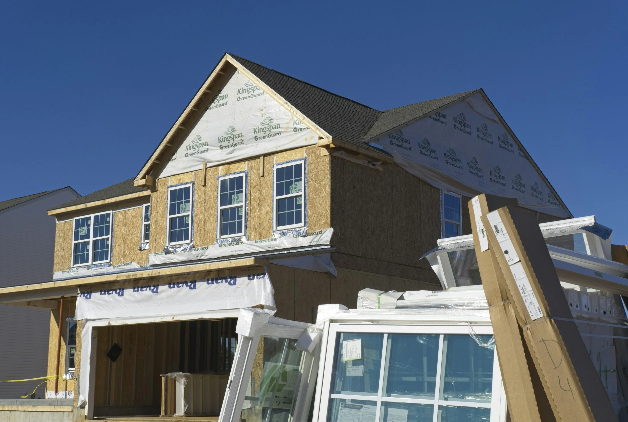 Construction site of a house with a partially built wooden frame, windows installed, and house wrap labeled Kingspan GreenGuard, with a clear blue sky in the background.