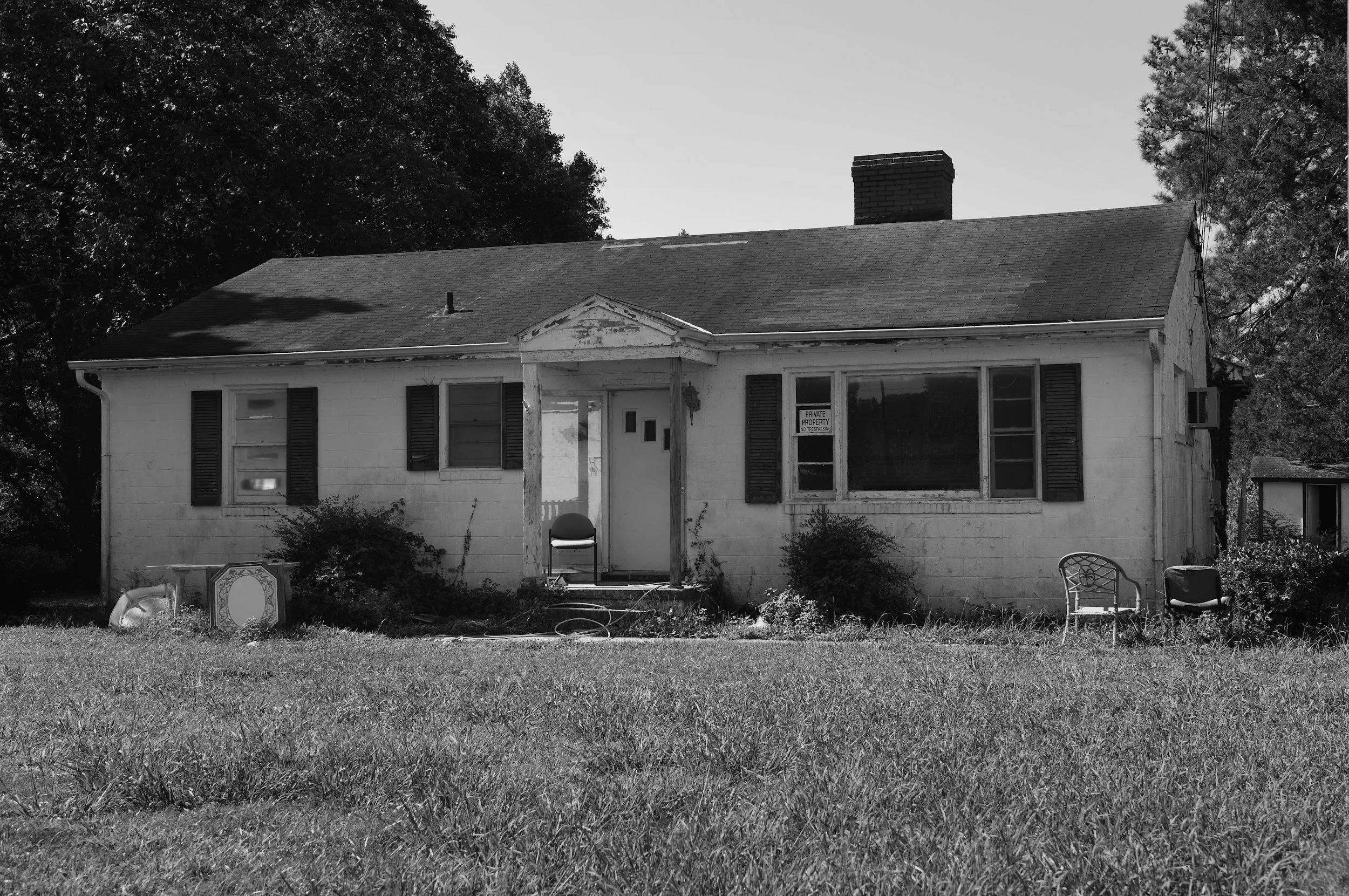 A black and white photo of an old house with a front porch, surrounded by a lawn and some outdoor furniture including chairs and a small table. Trees are in the background, and there is a 