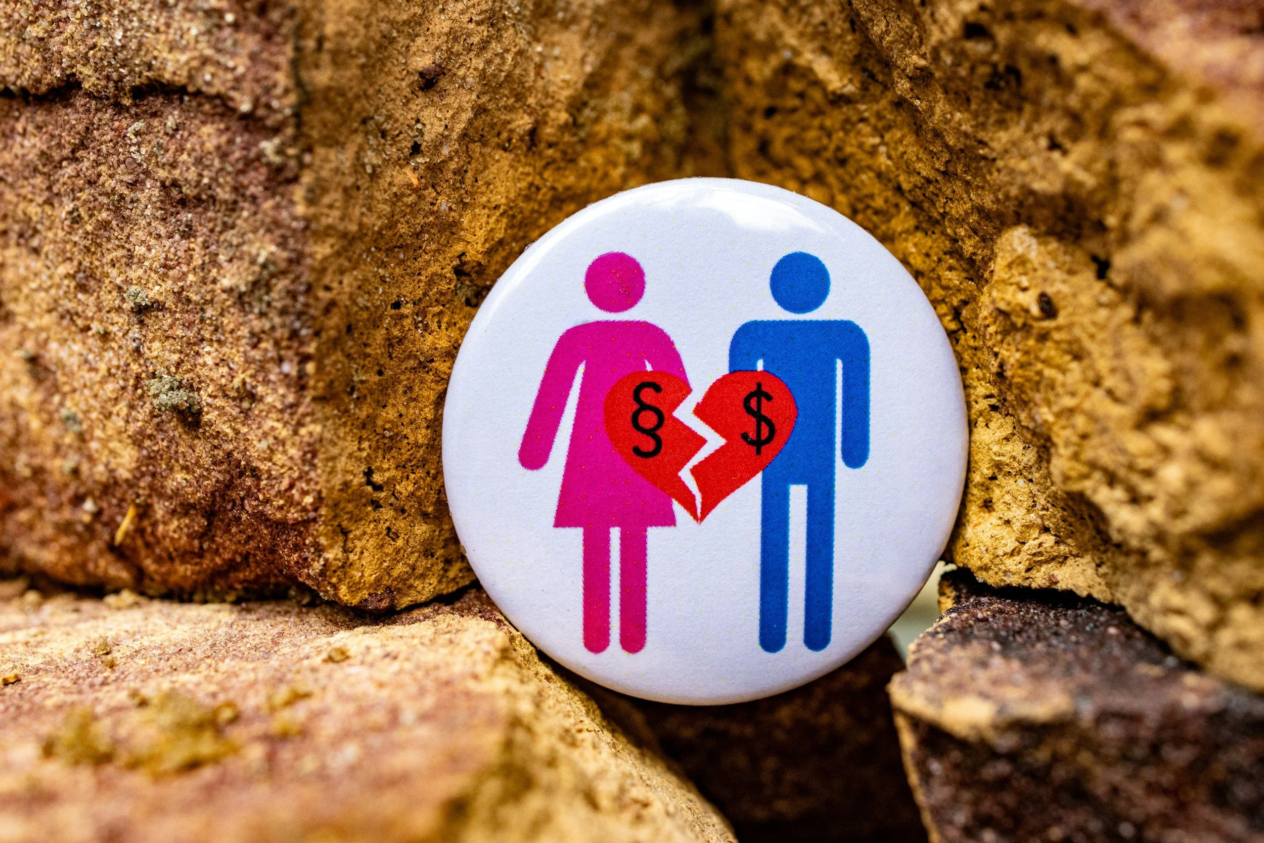 A pin with a pink female and blue male symbol, divided by a jagged line with dollar signs, placed among rocks or dirt.