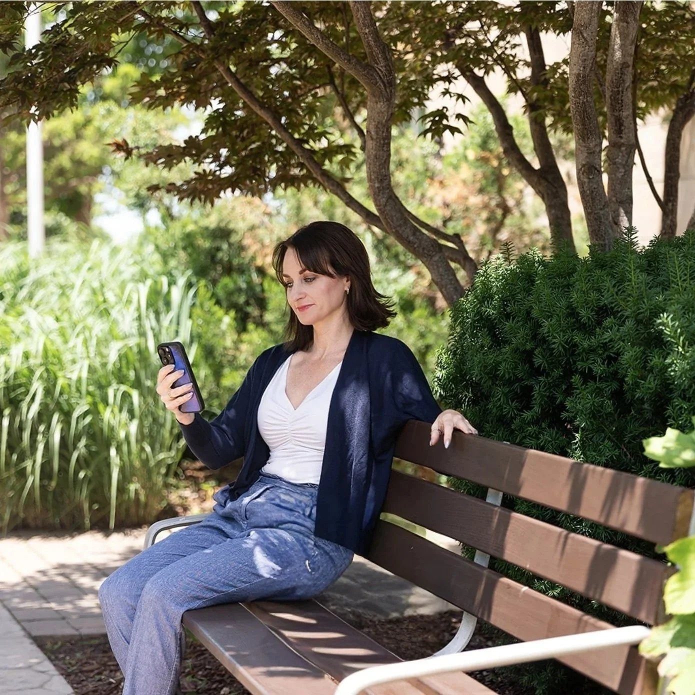 A woman sitting on a park bench, looking at her phone, surrounded by green foliage and trees.