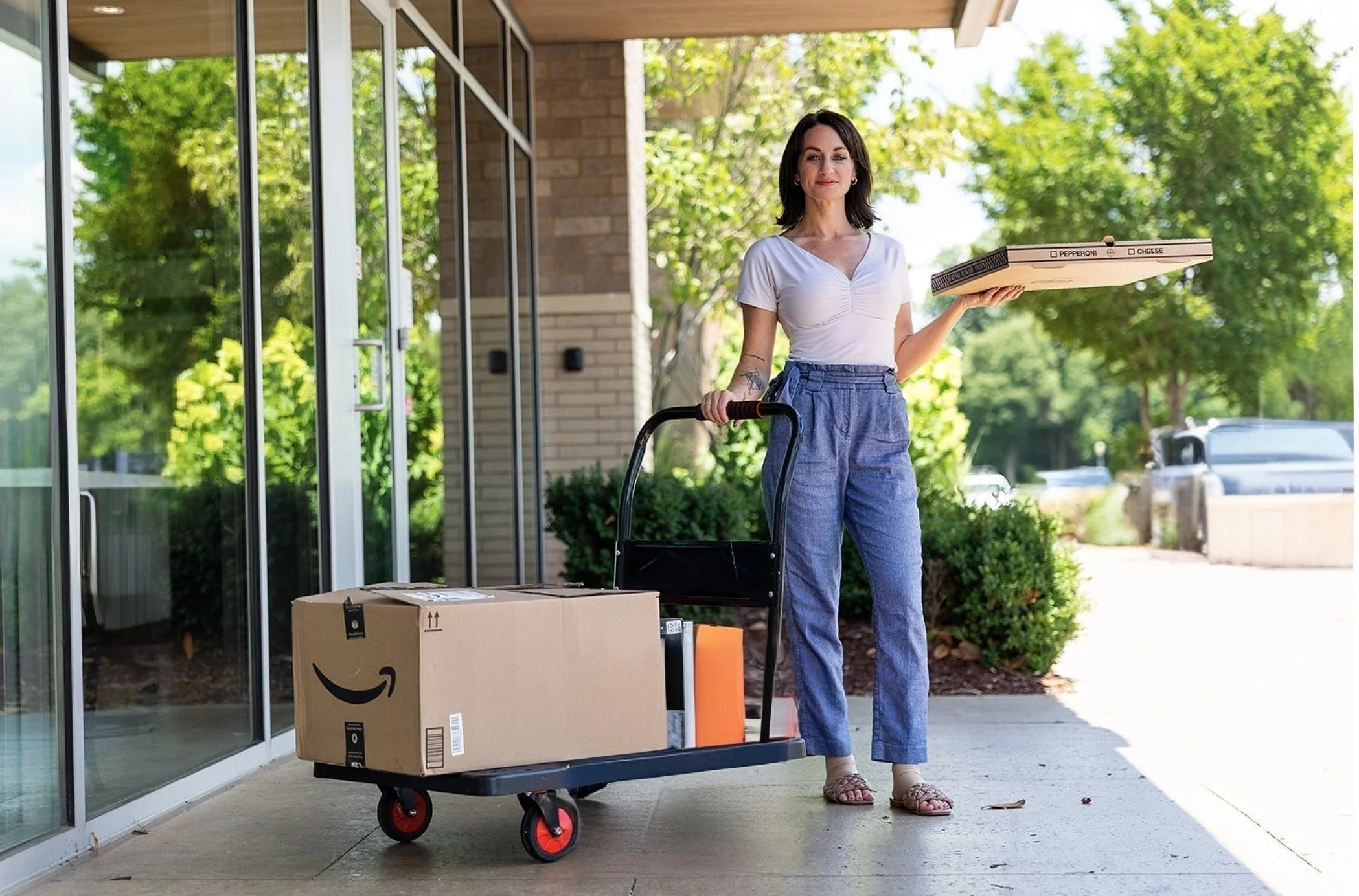 A woman with dark hair, wearing a white top and blue pants, holding a pizza box in one hand and pushing a cart with Amazon packages on it outside a building with glass doors and greenery in the background.