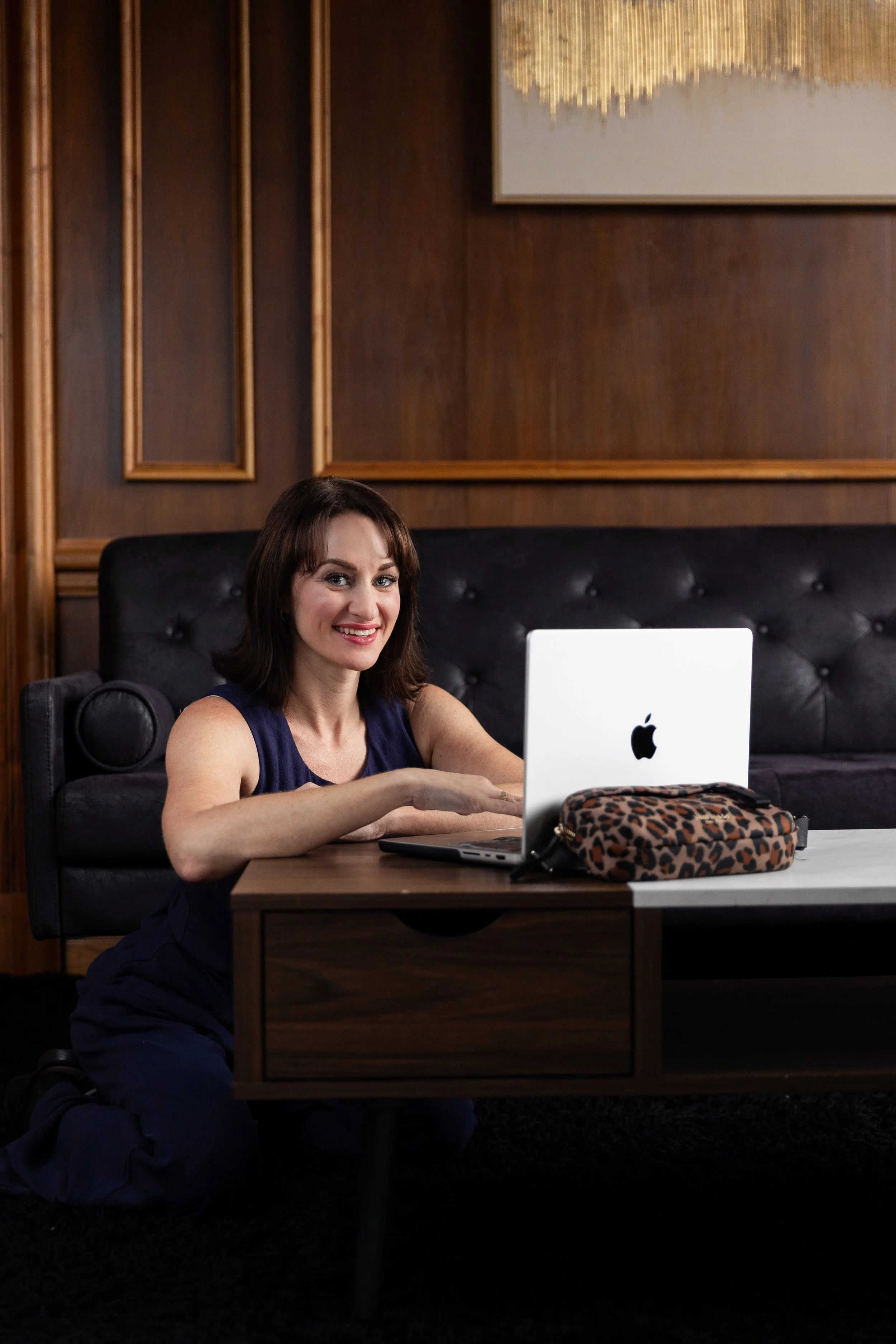 A woman with shoulder-length brown hair sitting at a desk, smiling at the camera, with an Apple iMac computer in front of her and a leopard-print bag on the desk. The background features dark wood walls with framed artwork.