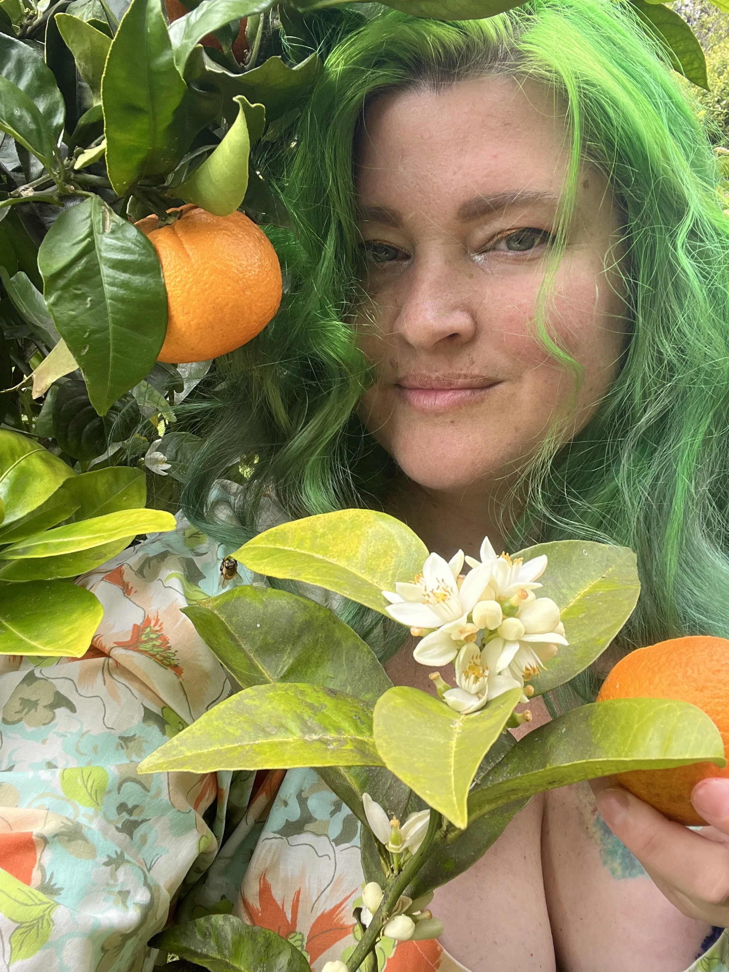 A woman with green hair, facial freckles, and a gentle smile is surrounded by lush green leaves, white flowers, and bright orange fruits in a natural outdoor setting.