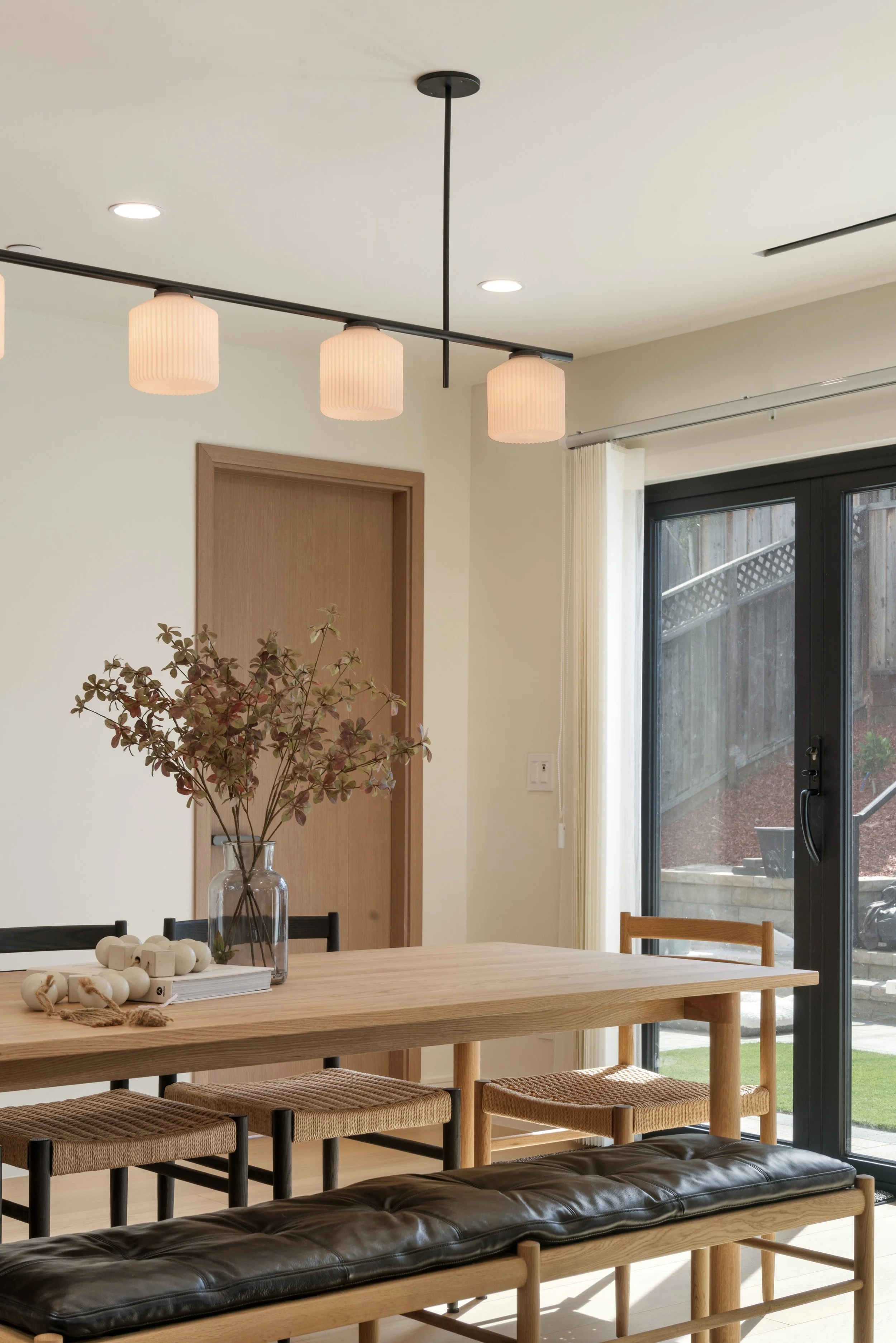 A dining area with a wooden table, black leather cushioned bench, and woven barstools. A tall glass vase with branches and some decorative objects are on the table. overhead lighting features three pinkish lounge-style lamps, and windows with white curtains and a sliding glass door lead to a patio with a wooden fence and outdoor furniture.