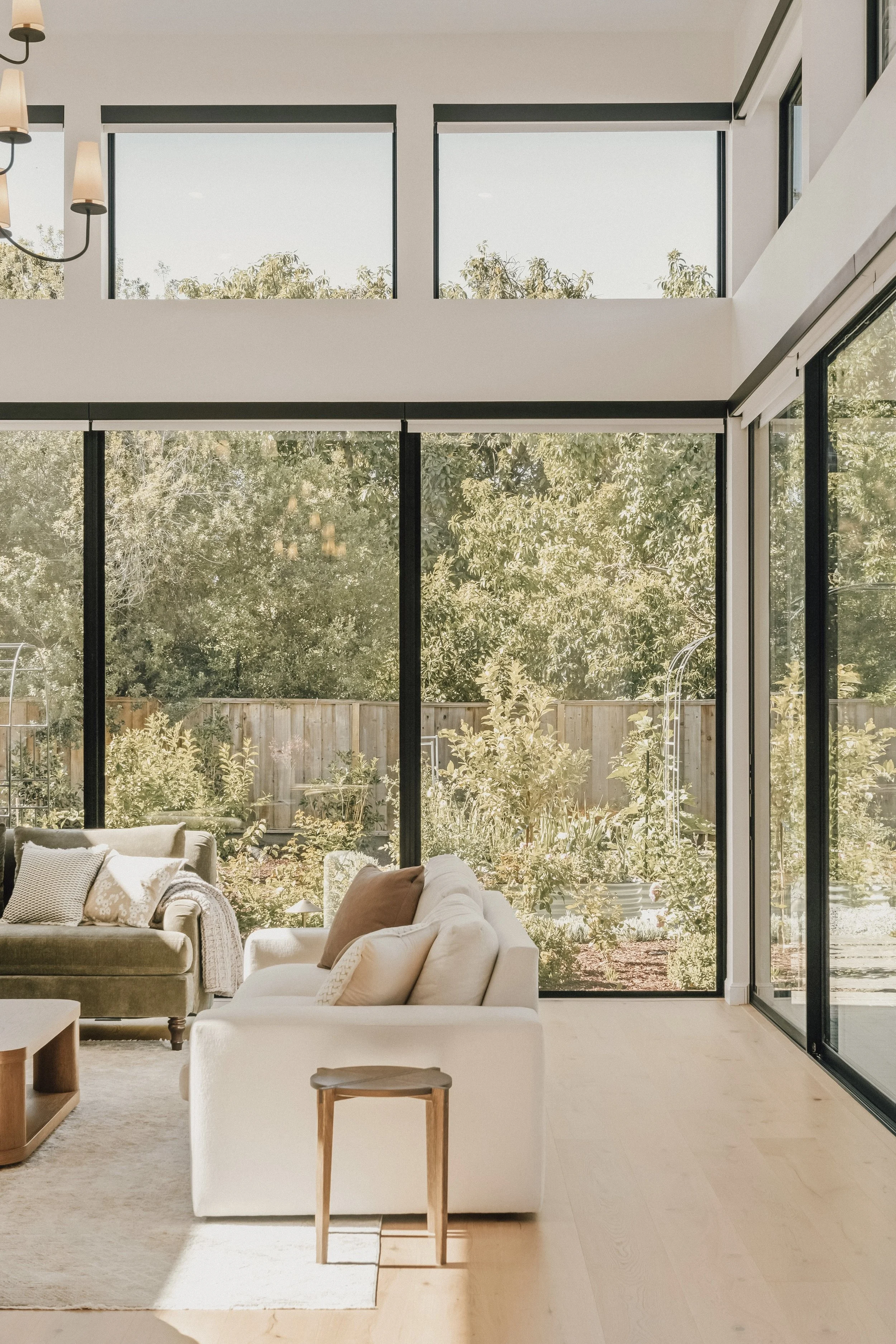 Living room with large windows, beige sofas with pillows, a wooden side table, and a view of a garden with trees and a wooden fence.