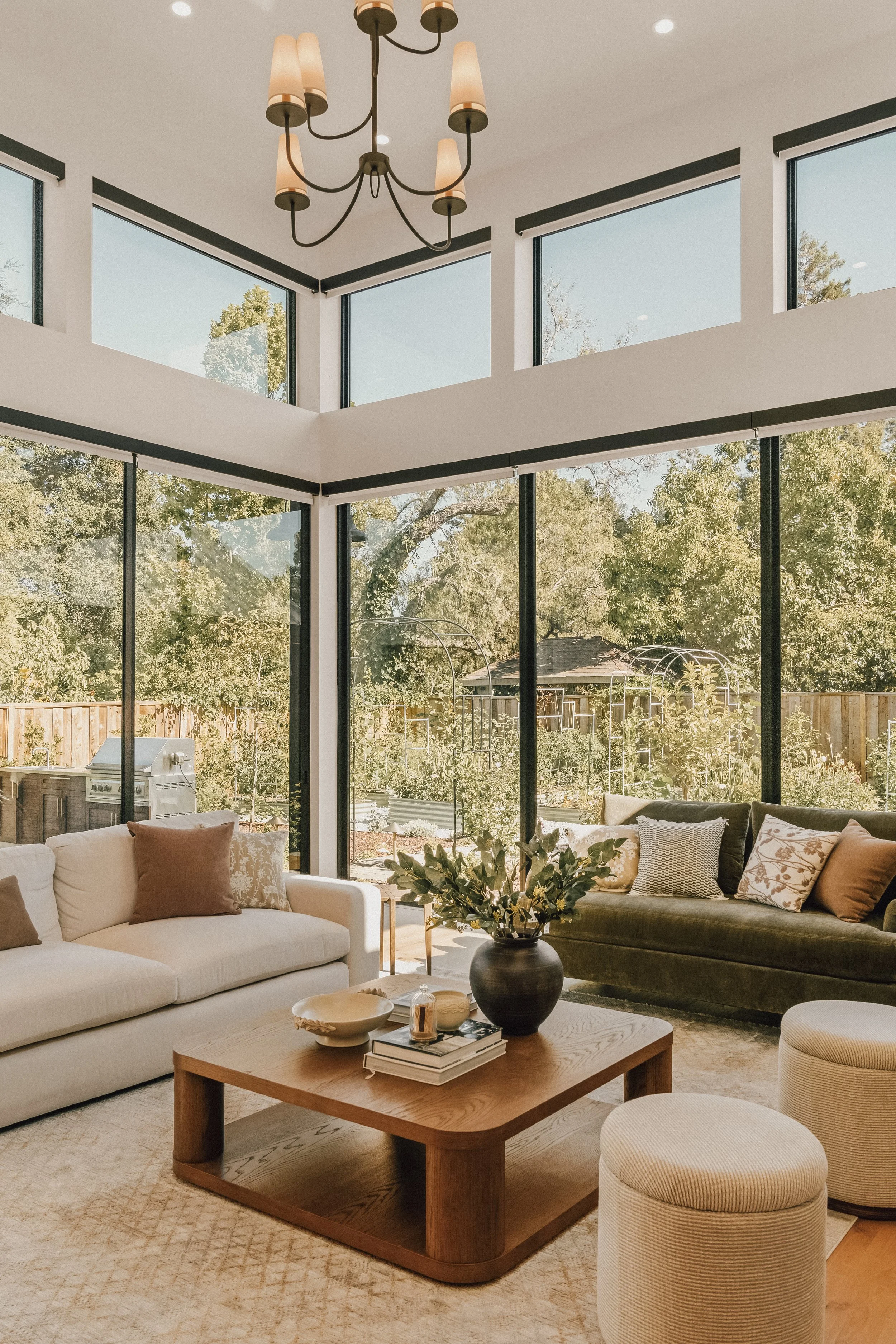 Living room with beige sofa, black sofa, round otteg, wooden coffee table with books and decorative objects, large window with trees outside, potted plant, chandelier with multiple lamps, ceiling with recessed lighting.