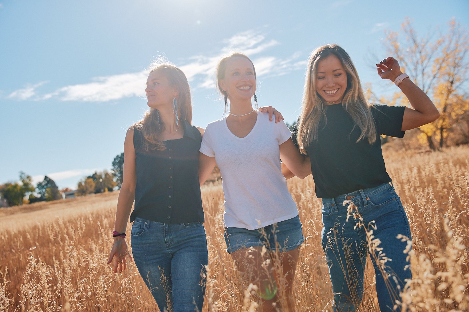 Three young women walking together in a golden field on a sunny day, smiling and enjoying each other's company.