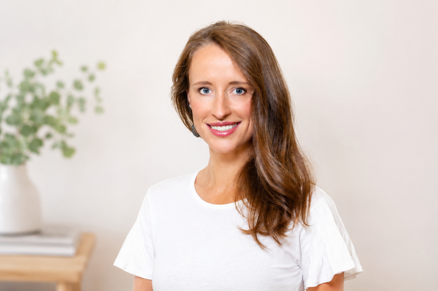 A smiling woman with long, wavy brown hair wearing a white t-shirt, standing in a minimalistic room with a potted plant on a wooden table in the background.
