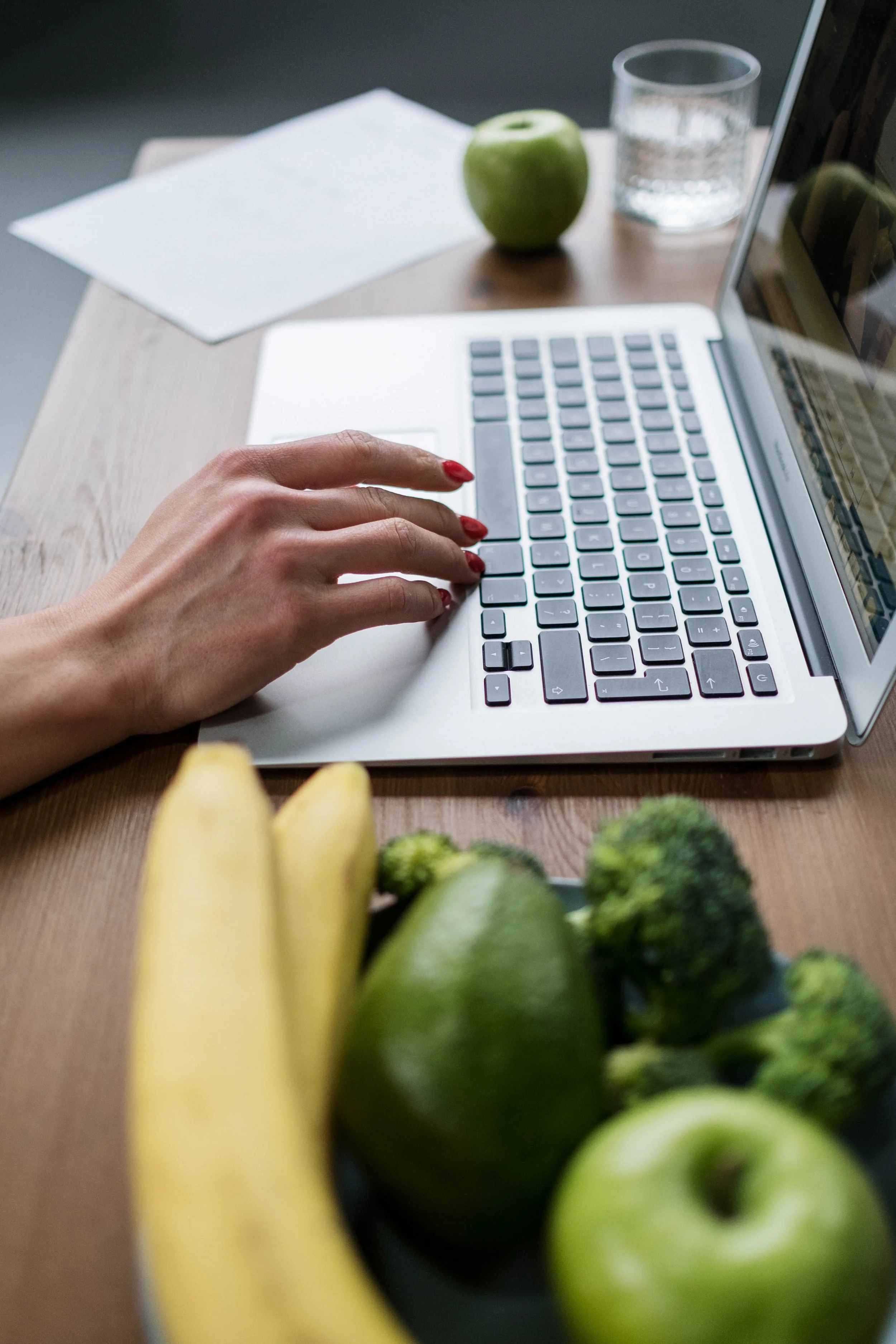 A person using a laptop at a desk with an apple, glass of water, bananas, broccoli, and avocados nearby.