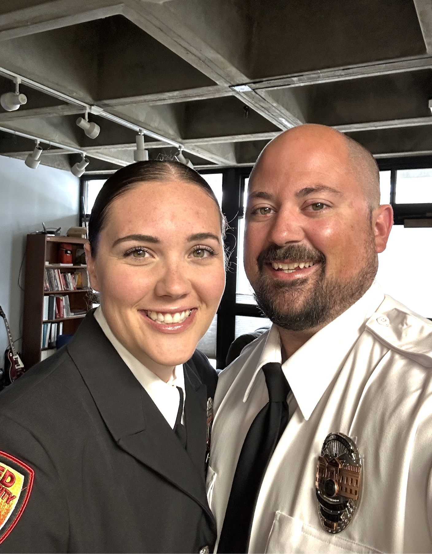A woman in a police uniform and a man in a security uniform smiling and taking a selfie inside an office with bookshelves and large windows.