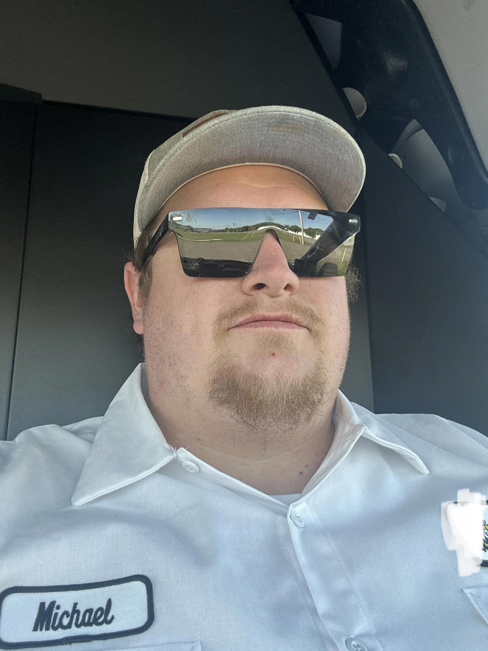 Close-up of a man with a beard and mustache wearing a white uniform shirt with a name tag that says 'Michael.' He has on a beige cap and reflective sunglasses showing a road and landscape. The image appears to be taken from the front seat of a vehicle.