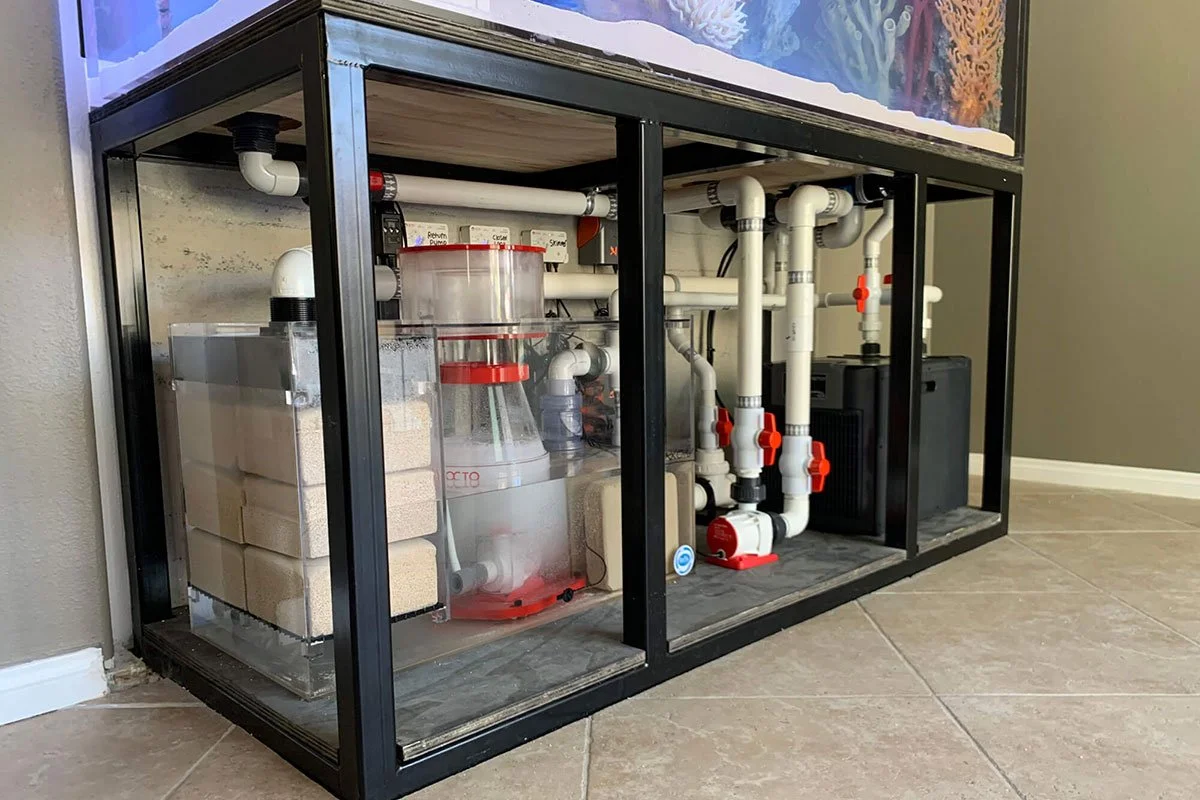 Aquarium filter system with white PVC pipes, a water pump, filter media, and a heater unit, set up under an aquarium on a black metal frame.