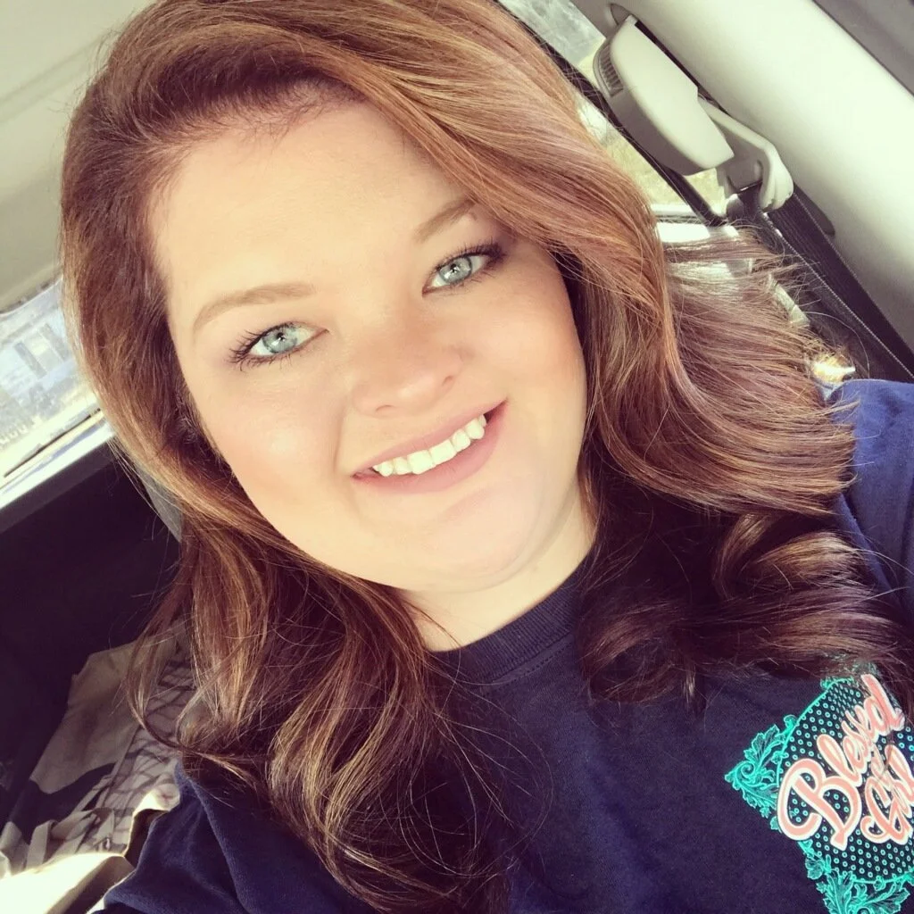 A young woman with red hair and blue eyes smiling inside a car.