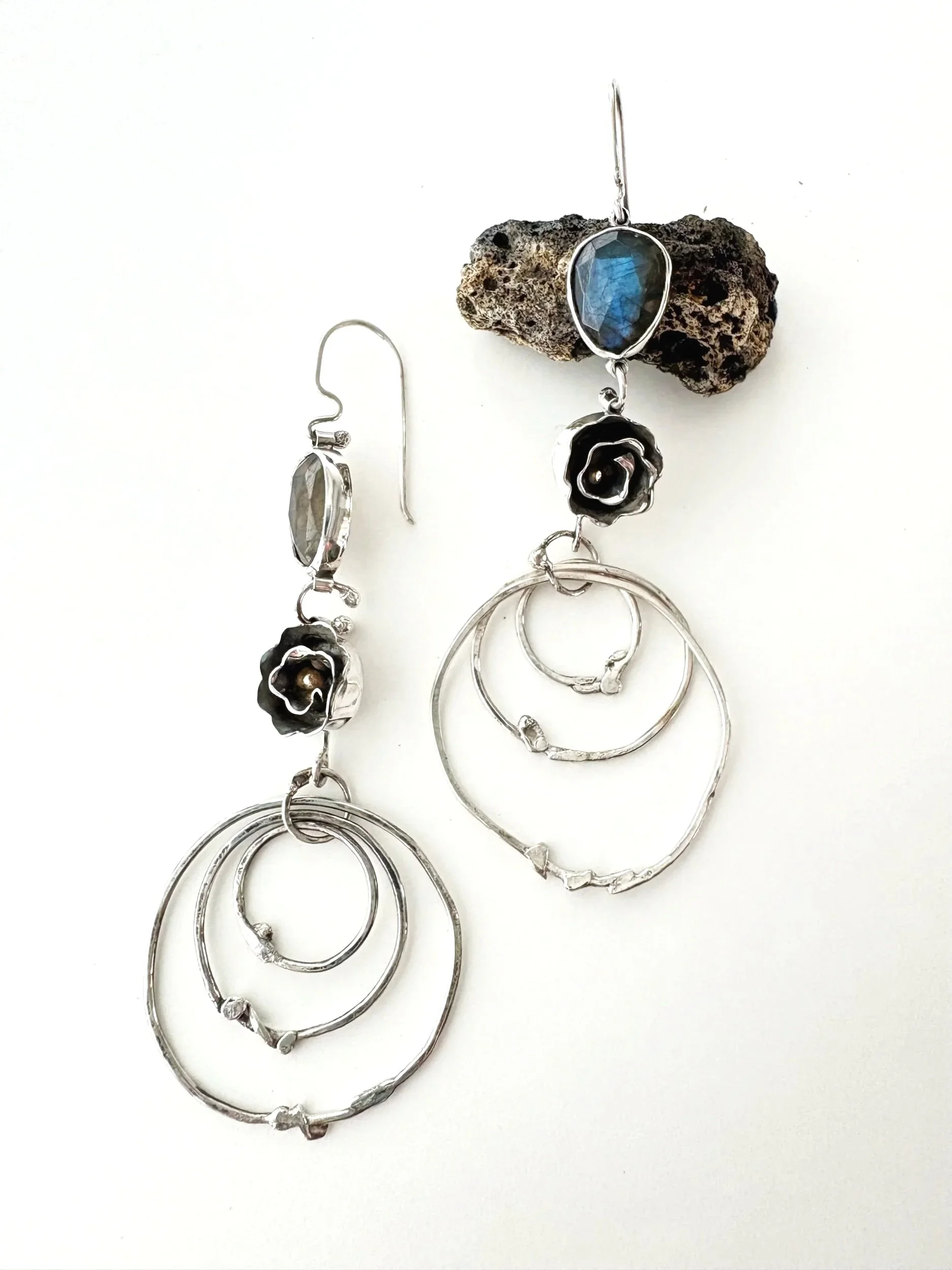 Silver earrings and bangles with black roses and blue gemstones, placed on a white background with a textured rock.