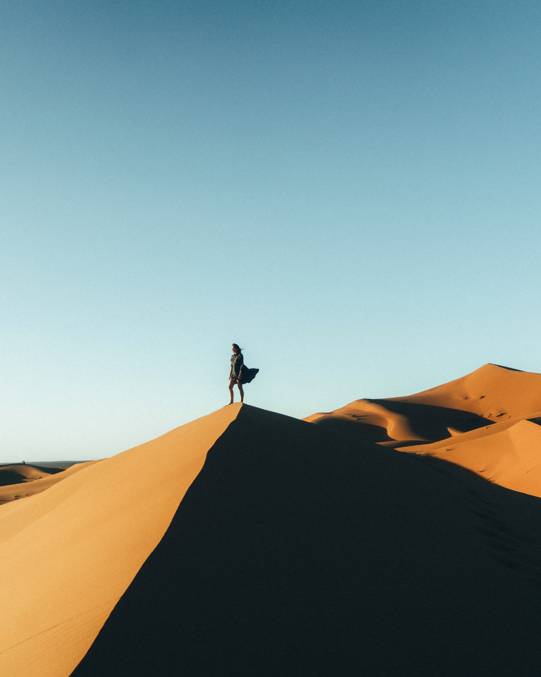 A person standing on a sand dune in a desert landscape under a clear blue sky.