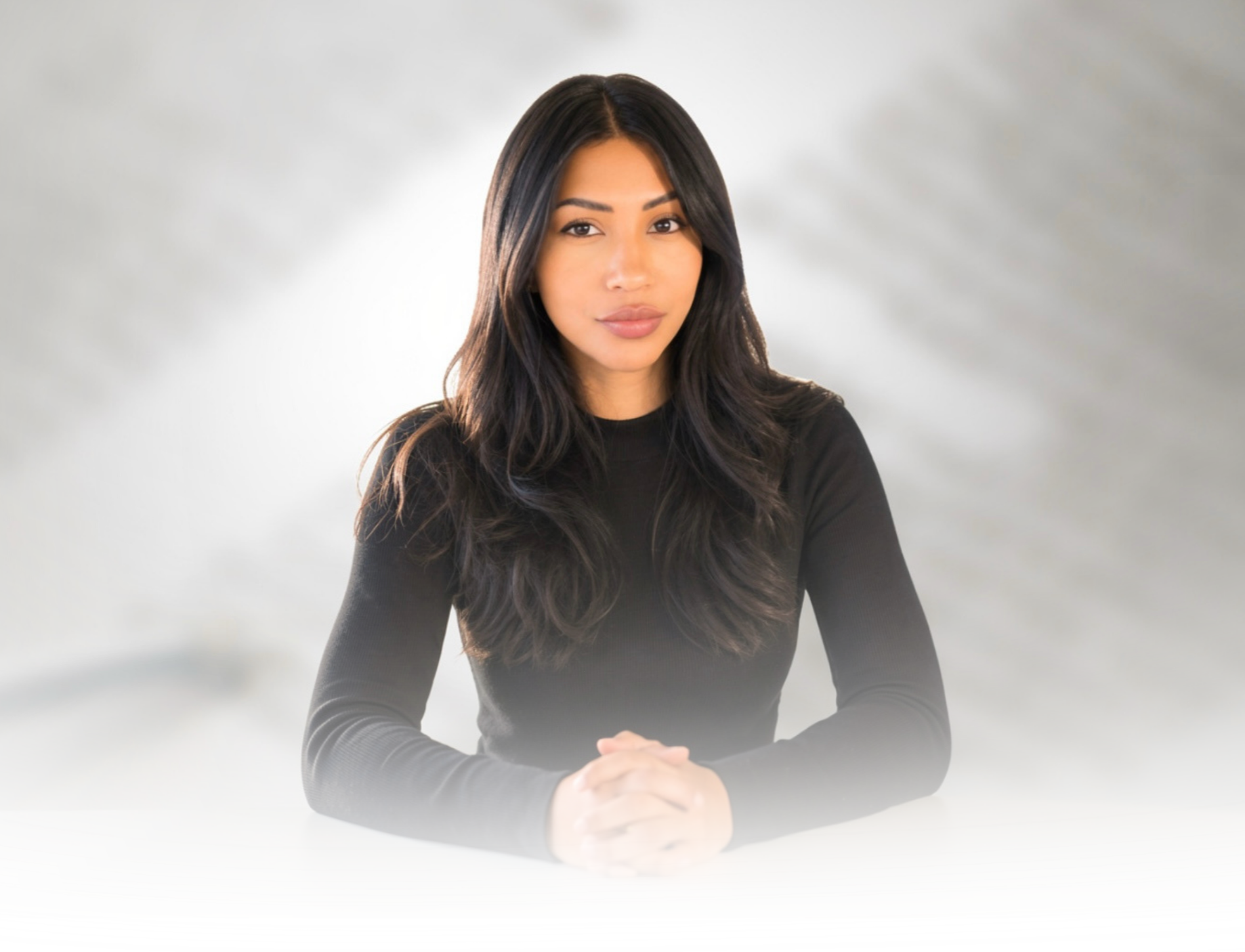 A woman with long, dark hair and light makeup sitting at a white table with her hands clasped, wearing a black long-sleeve top, against a blurred light gray background.