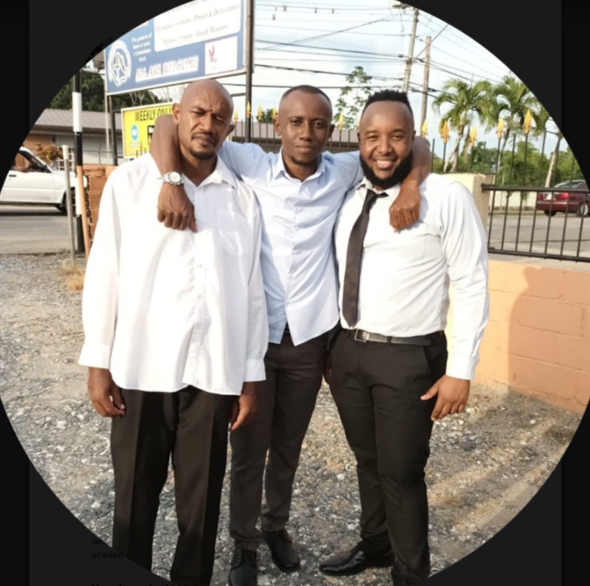 Three men in formal attire standing outdoors. They have their arms around each other's shoulders, smiling at the camera. The background shows a parking lot, power lines, trees, and a signboard.