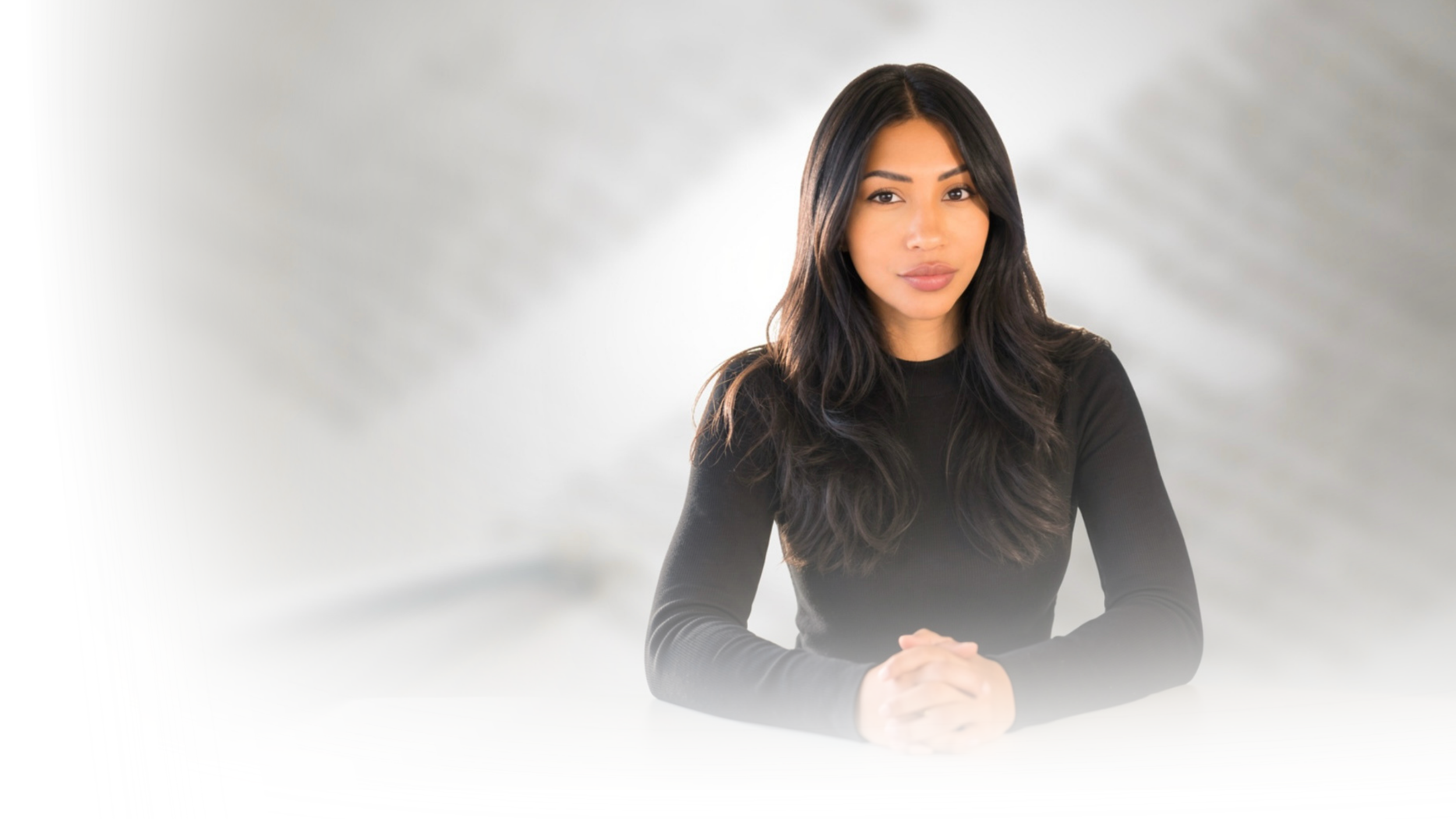 A woman with long dark hair and wearing a black long-sleeve shirt, sitting at a table with hands clasped, against a soft gray background.