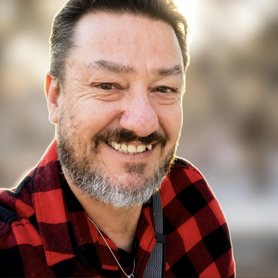 Close-up of a man smiling outdoors with sunlight in the background, wearing a red and black checkered shirt.