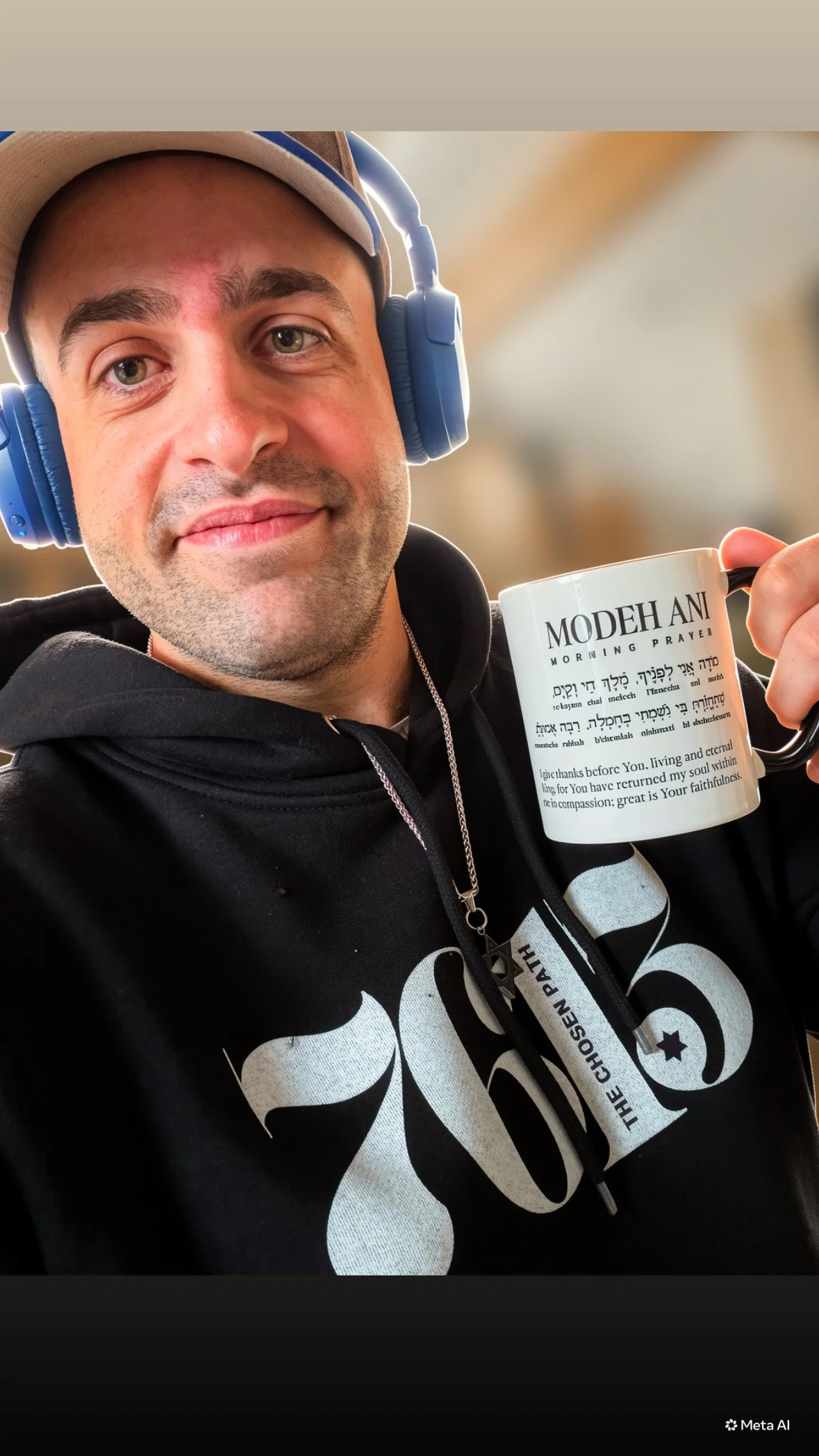 A man with green eyes and a light beard wearing a beige cap and blue headphones is smiling and holding a white mug with black text, wearing a black hoodie with a white design.