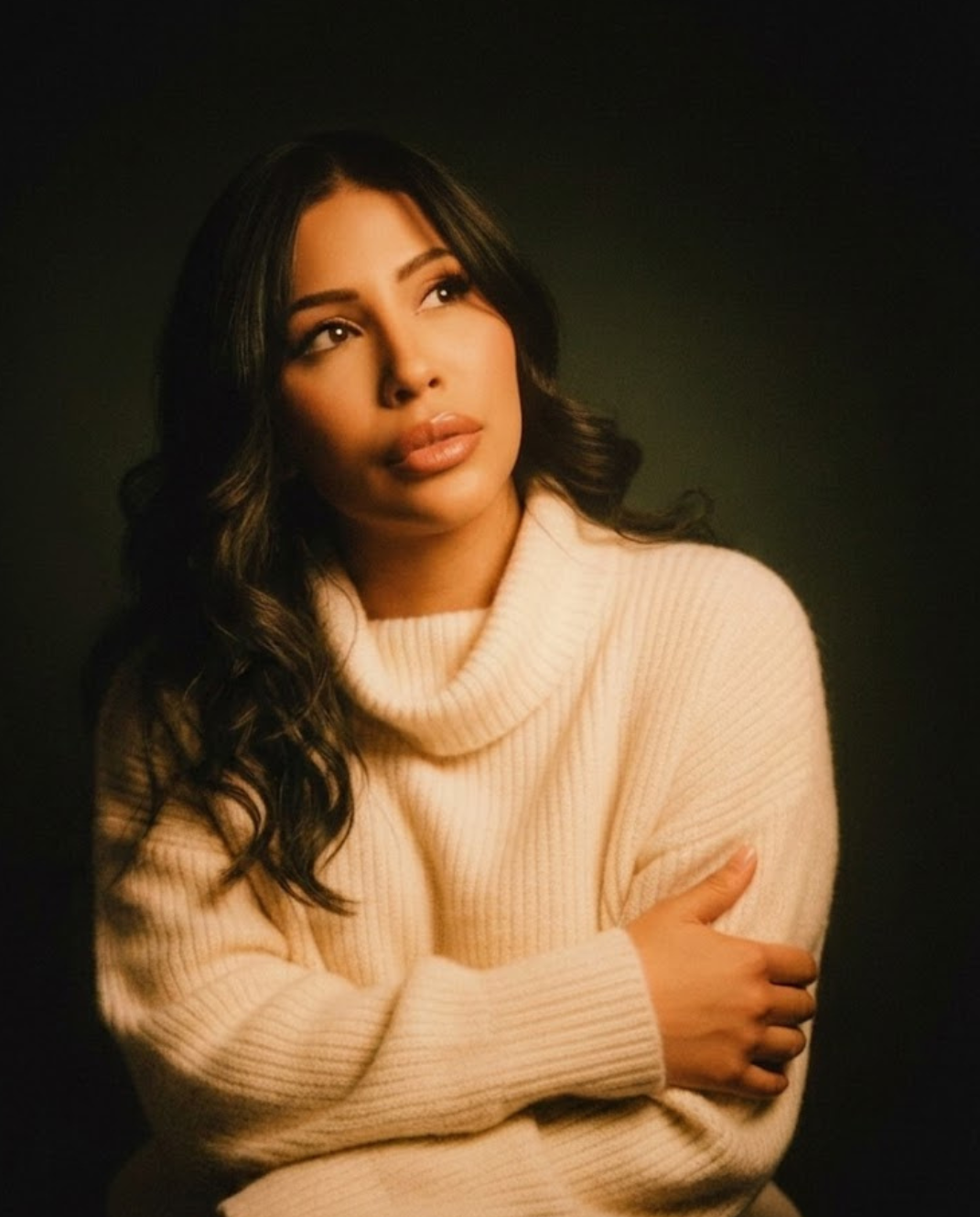 A woman with long dark wavy hair wearing a cream-colored turtleneck sweater, looking slightly upwards with her arms crossed.