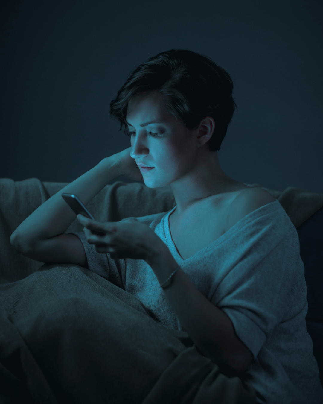 A woman with short dark hair sitting on a couch at night, looking at her smartphone with a focused expression, illuminated by the blue light from the device.