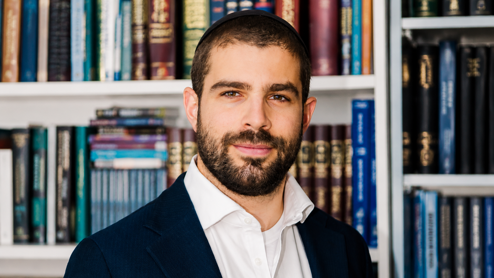 Rabbi Noah Tile with a beard and short hair wearing a white shirt and dark blazer in front of bookshelves.