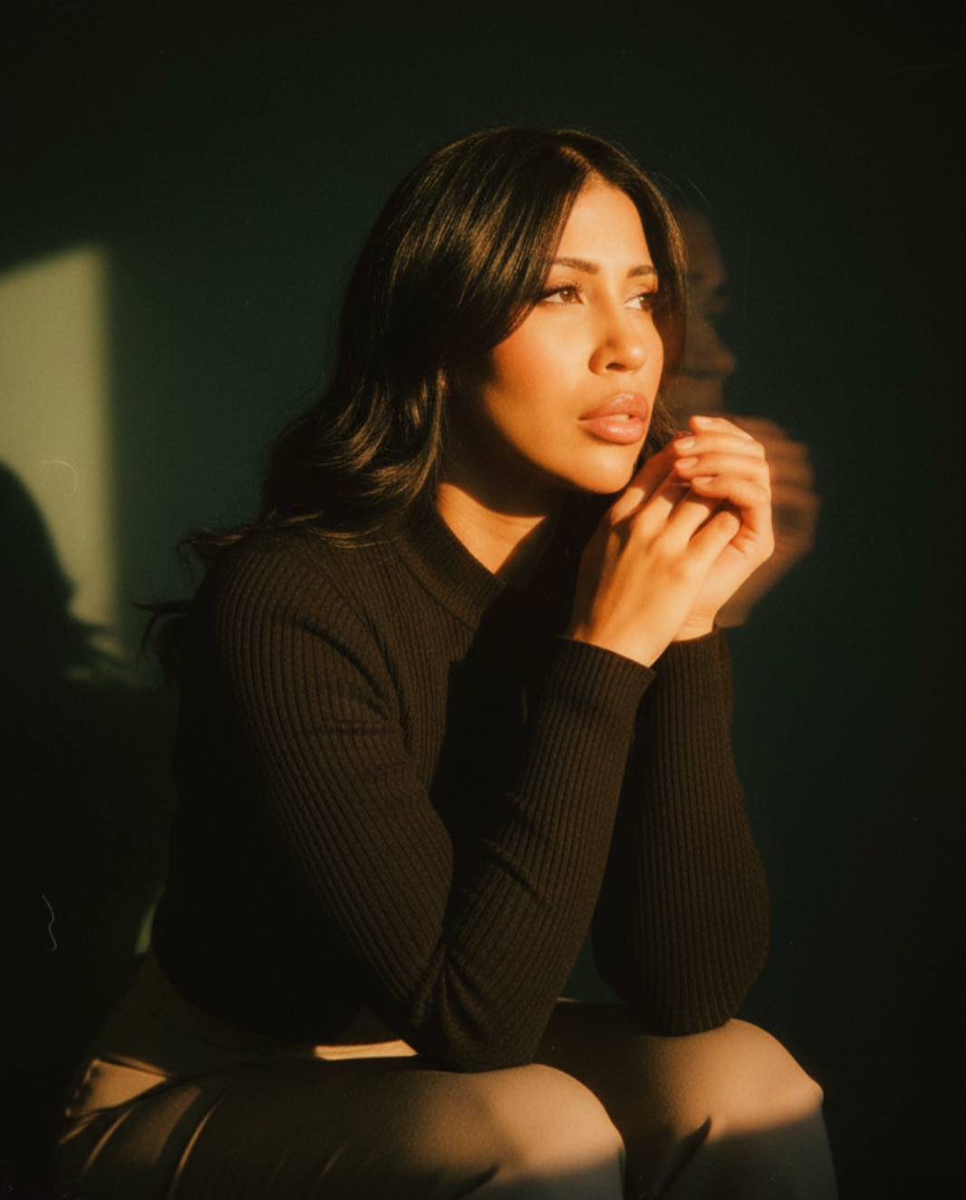 Tassja Cadoch with dark hair looking contemplative, sitting with her hands clasped near her face, lit by soft, warm lighting.