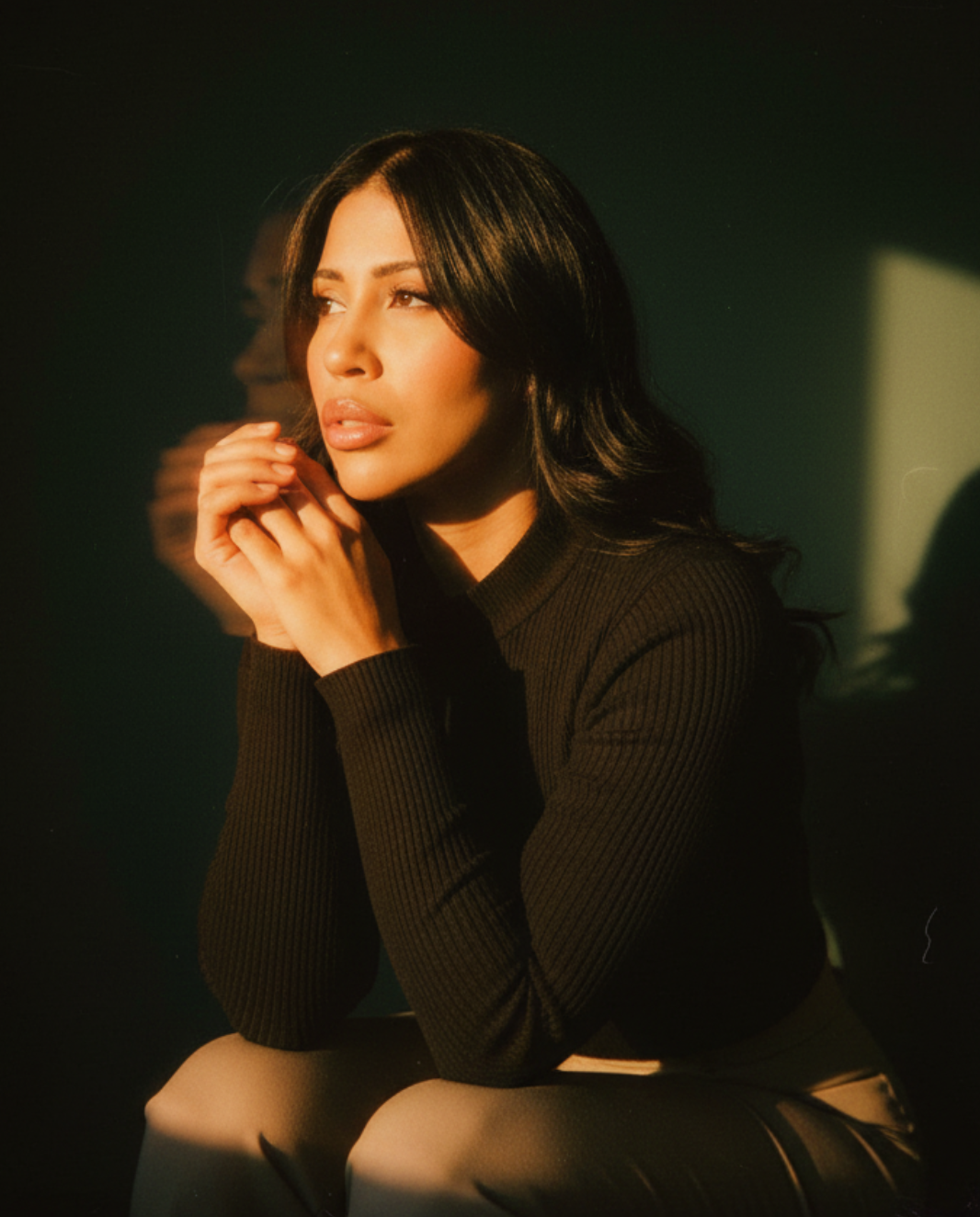 A woman with dark hair and a black ribbed top sitting with her hands clasped near her face, lit with warm light and a dark background.