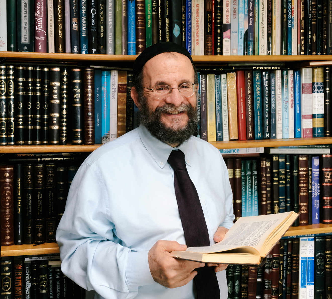A smiling man with glasses, a beard, wearing a white shirt, dark tie, and a black kippah, holding an open book in front of a bookshelf filled with colorful books.