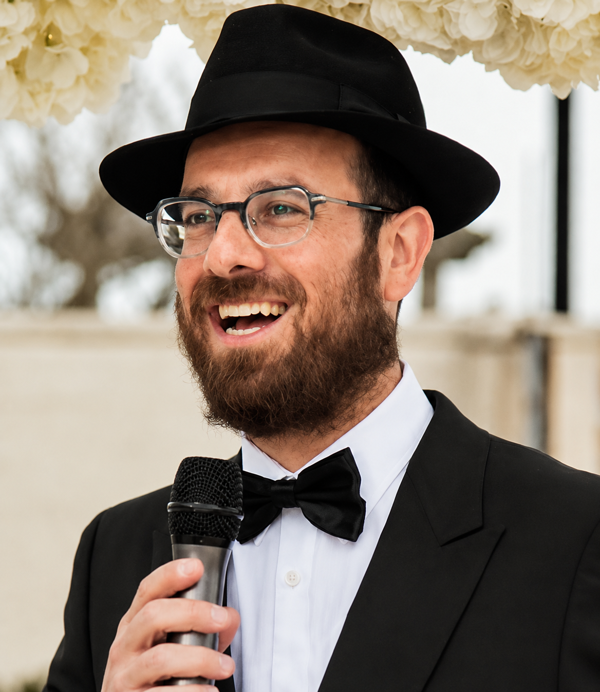 Rabbi Raps - Rabbi Avrohom Rapoport in a tuxedo and glasses, wearing a black fedora, smiling and holding a microphone, speaking at an event with white flowers in the background.